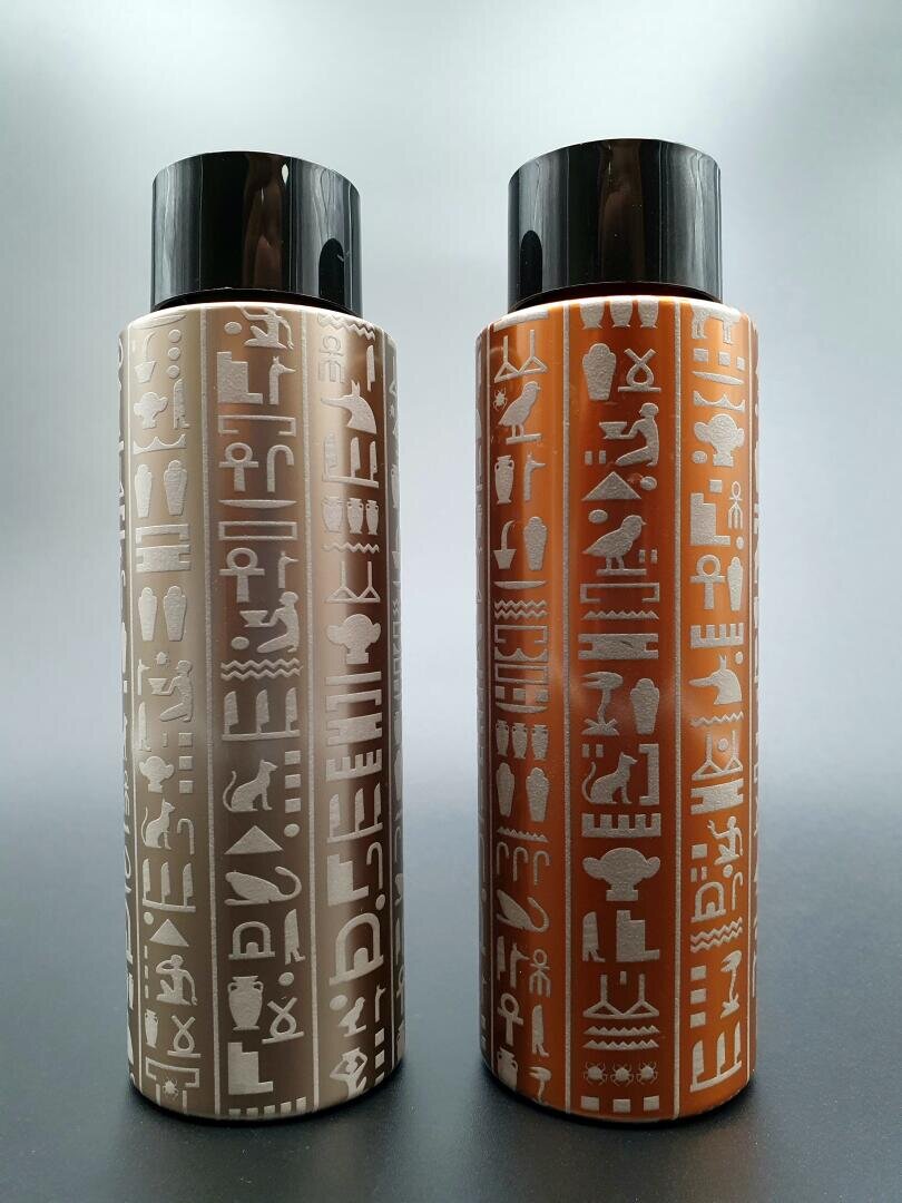 Zwei zylindrische Flaschen mit schwarzen Kappen, verziert mit vertikalen Reihen ägyptischer Hieroglyphen. Eine Flasche ist beige, die andere rötlich-braun, beide stehen aufrecht auf einem neutralen grauen Hintergrund.
