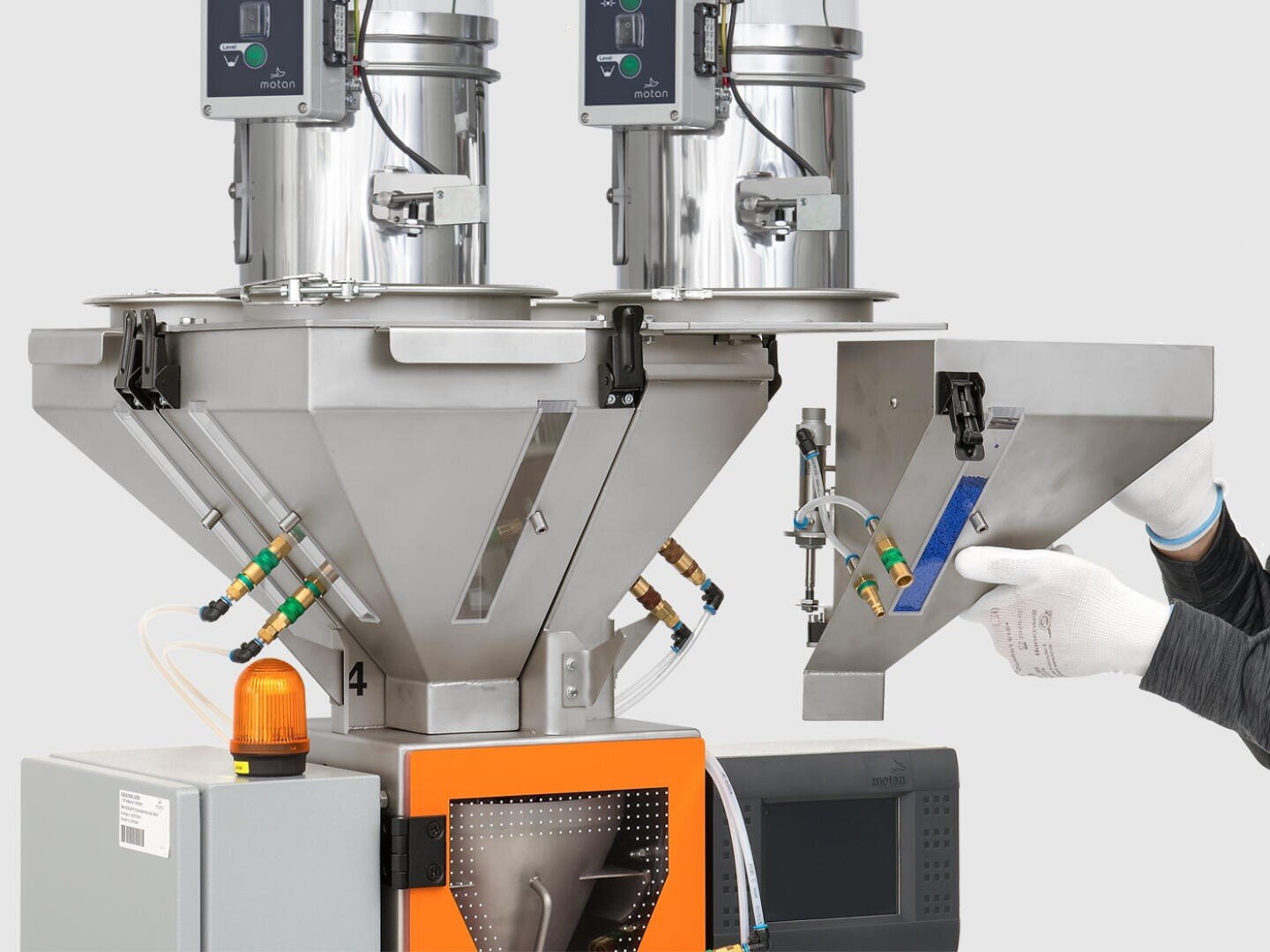 Une personne portant des gants blancs ajuste une pièce métallique sur une machine industrielle avec deux unités cylindriques sur le dessus et des panneaux de commande ; un petit voyant d'avertissement orange est visible sur le devant.