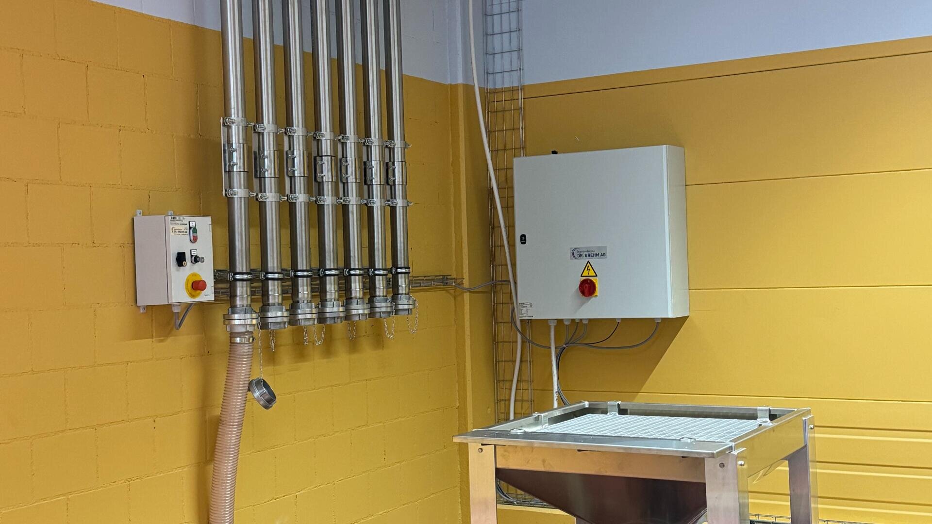 Ein Industrieraum mit gelben Wänden, Metallrohren, Schalttafeln und einer Maschine oder einem Trichter aus Edelstahl in der Ecke. Der Raum wirkt sauber und aufgeräumt.