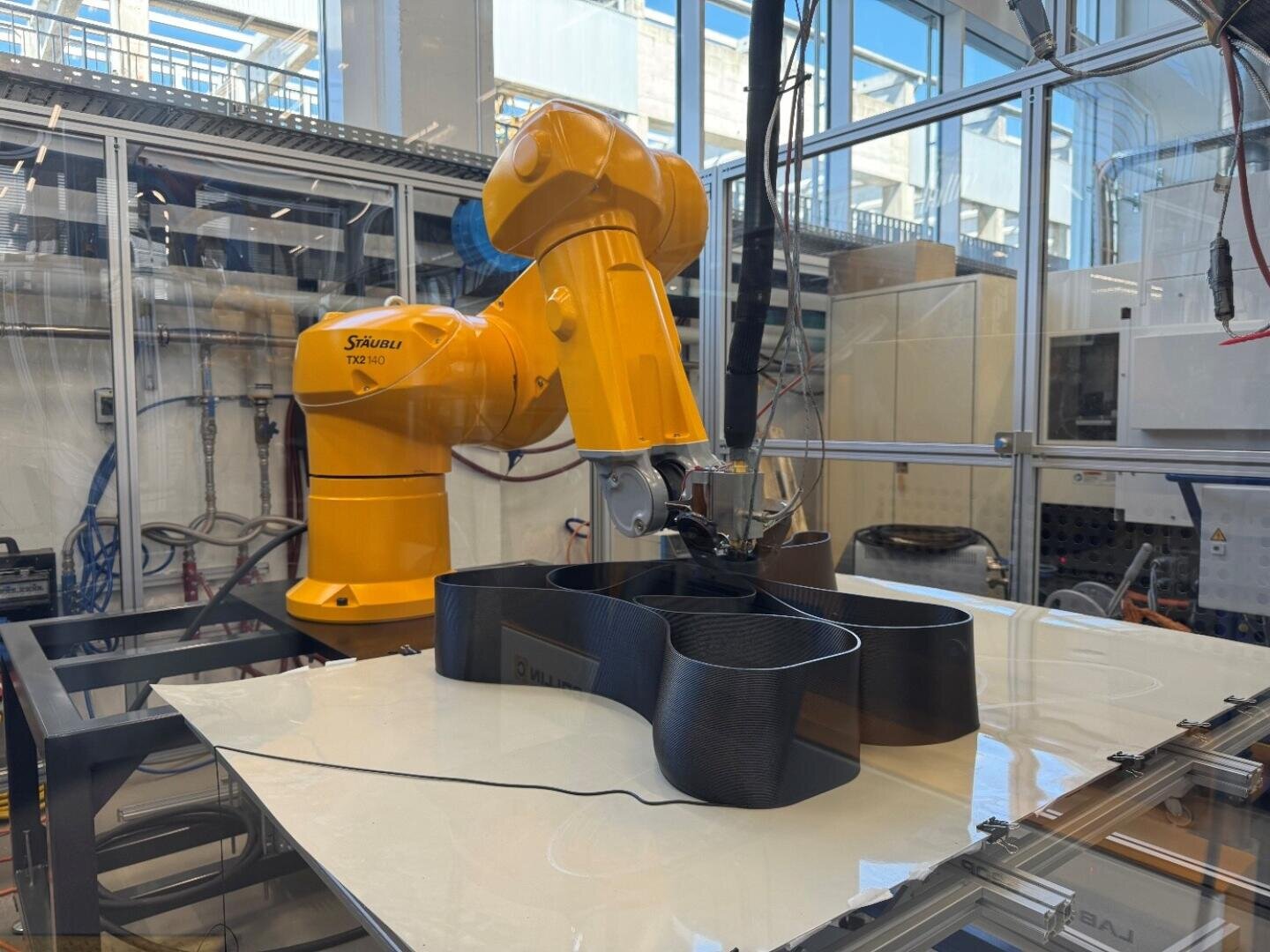 Ein gelber Industrieroboterarm druckt in einem Labor mit Maschinen, Glaswänden und sichtbaren Kabeln ein großes, schwarzes, wellenförmiges Objekt in 3D.