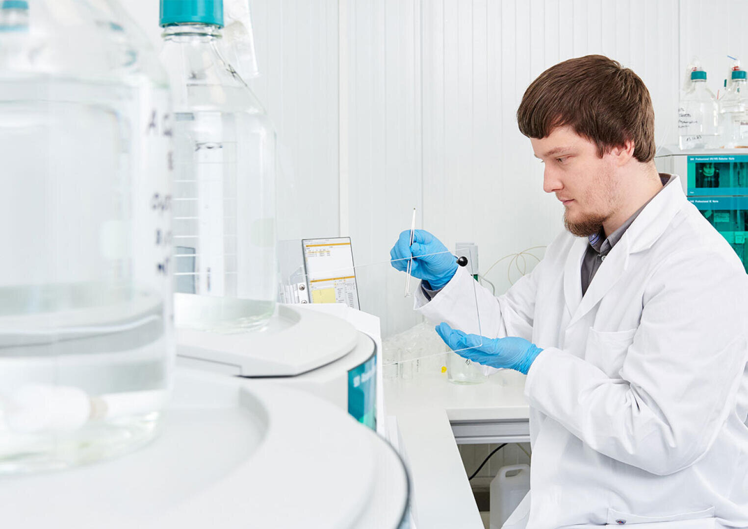 Ein Wissenschaftler in weißem Laborkittel und blauen Handschuhen arbeitet mit Laborgeräten, hält eine Pipette und einen kleinen Behälter in einer hellen, modernen Laborumgebung mit großen Glasflaschen im Vordergrund.