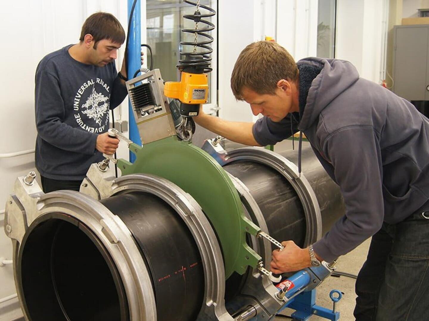 Zwei Männer bedienen Maschinen, um große schwarze Kunststoffrohre in einer Industriehalle auszurichten und zu verbinden. Ein Mann stellt die Ausrüstung ein, während der andere den Vorgang überwacht. Um sie herum sind Werkzeuge und mechanische Teile zu sehen.