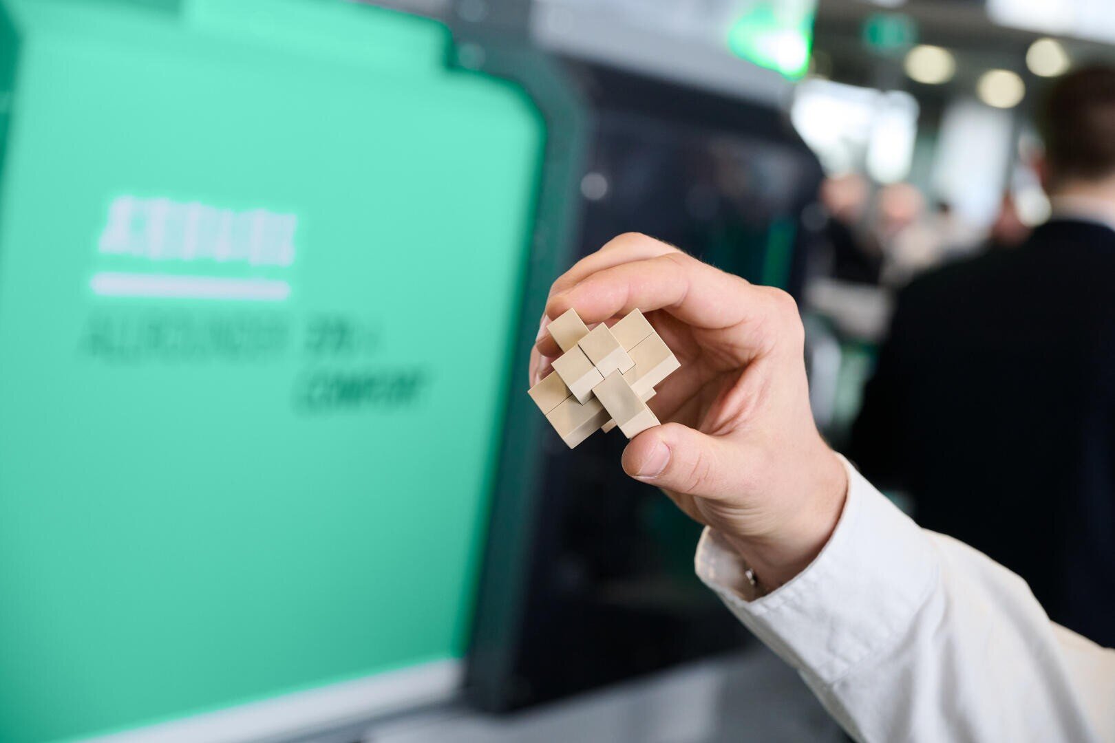 Une personne tient dans sa main une petite pièce de puzzle en bois en 3D, sur un fond flou vert et blanc avec du texte et des personnes dans un environnement intérieur.