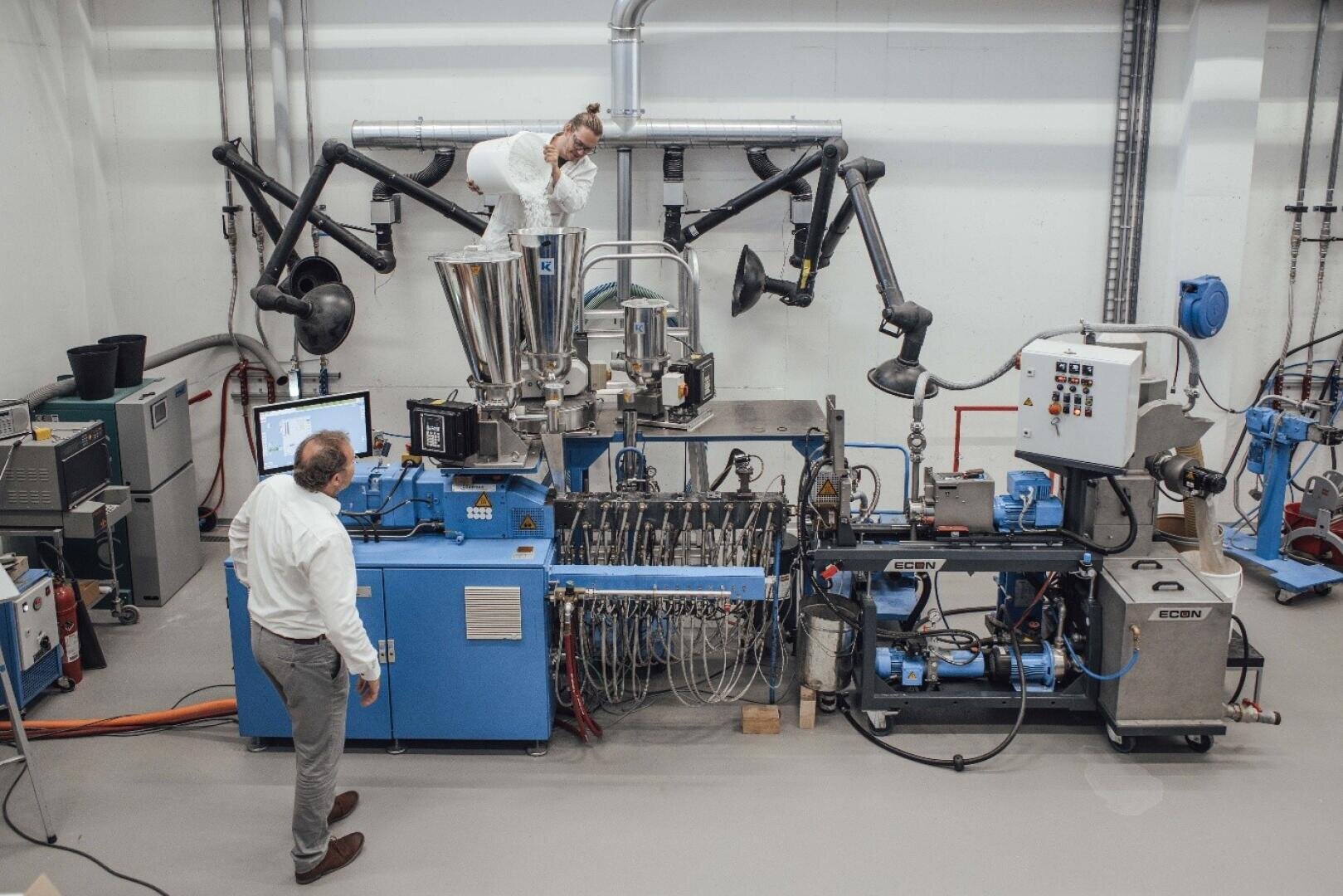 Zwei Personen in einem Labor mit Industriemaschinen; eine steht an einem Bedienfeld, während die andere einen Trichter über der Maschine inspiziert. Metallrohre und verschiedene Geräte umgeben sie in dem sauberen, modernen Arbeitsbereich.