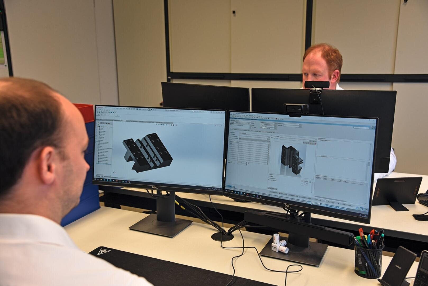 Zwei Personen arbeiten an Schreibtischen mit zwei Monitoren, auf denen 3D-Modellierungs- und technische Software angezeigt wird. Eine Person ist von der Kamera abgewandt, die andere ist teilweise hinter einem Monitor zu sehen. Auf dem Schreibtisch liegen Büromaterialien.