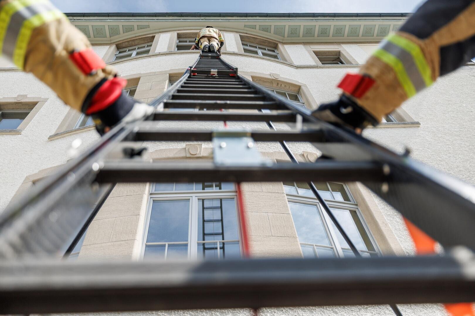 Une vue à faible hauteur de pompiers grimpant une échelle adossée à un bâtiment, l'un d'entre eux se tenant en haut, près d'une fenêtre supérieure, et un autre s'accrochant en bas, sur les côtés de l'échelle.