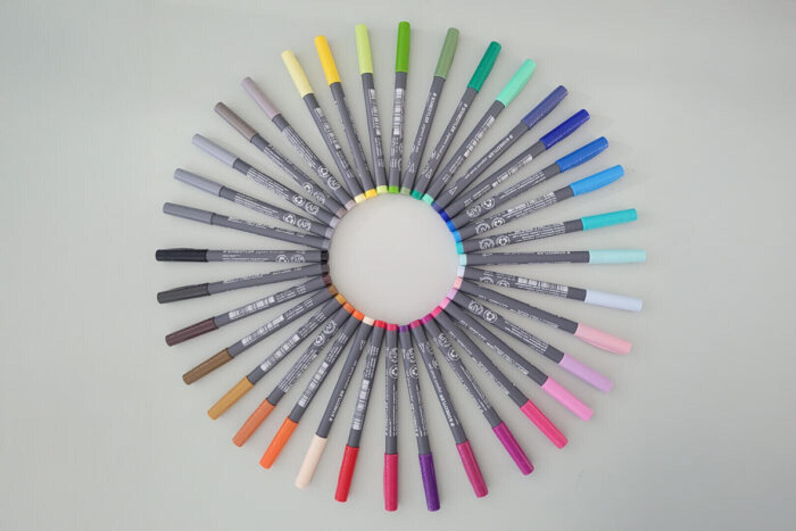Ein Kreis, der aus 36 farbigen Stiften besteht, die in einem Regenbogenverlauf angeordnet sind, wobei die Stiftspitzen nach außen zeigen und die Stiftkörper einen Ring um einen leeren Mittelpunkt auf einem hellen Hintergrund bilden.