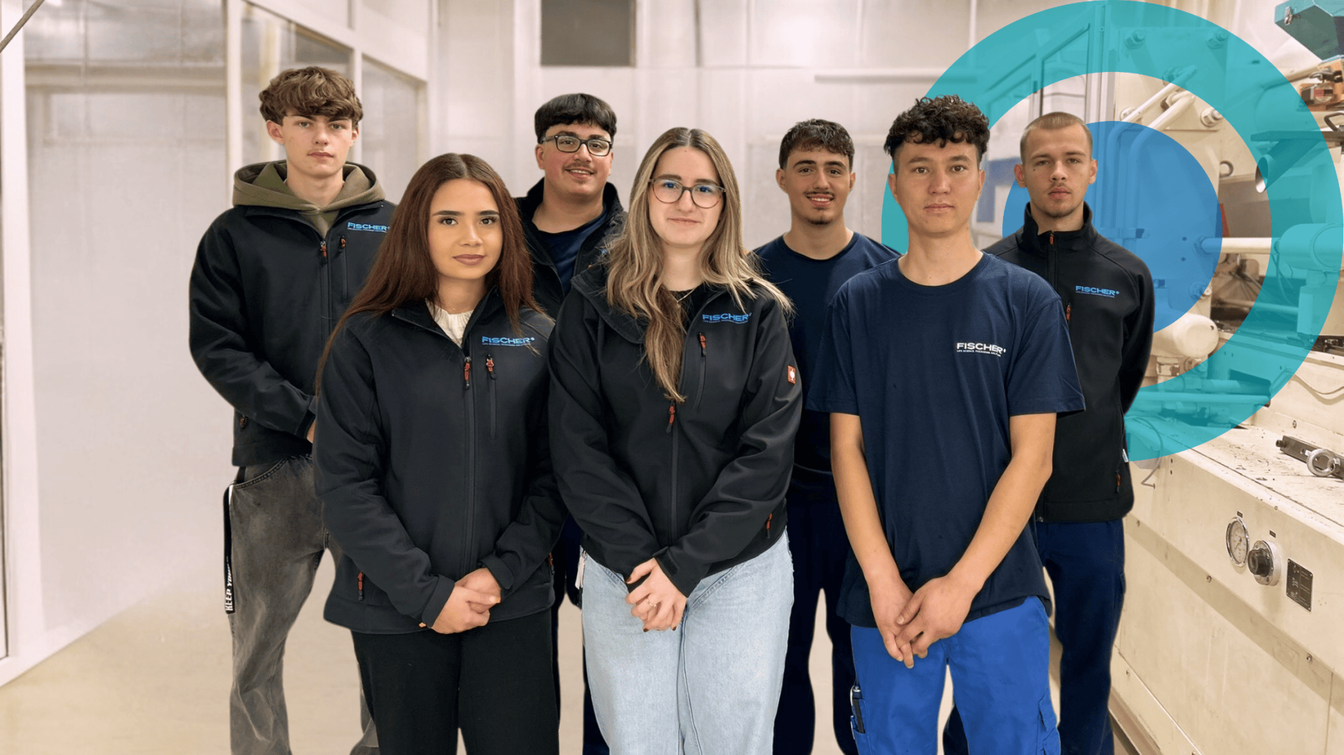 Sieben junge Erwachsene, vier Männer und drei Frauen, stehen in einem Innenraum zusammen. Die meisten tragen schwarze Jacken mit blauen Akzenten, zwei tragen marineblaue Hemden. Sie posieren und lächeln in einem hellen, industriell wirkenden Raum.