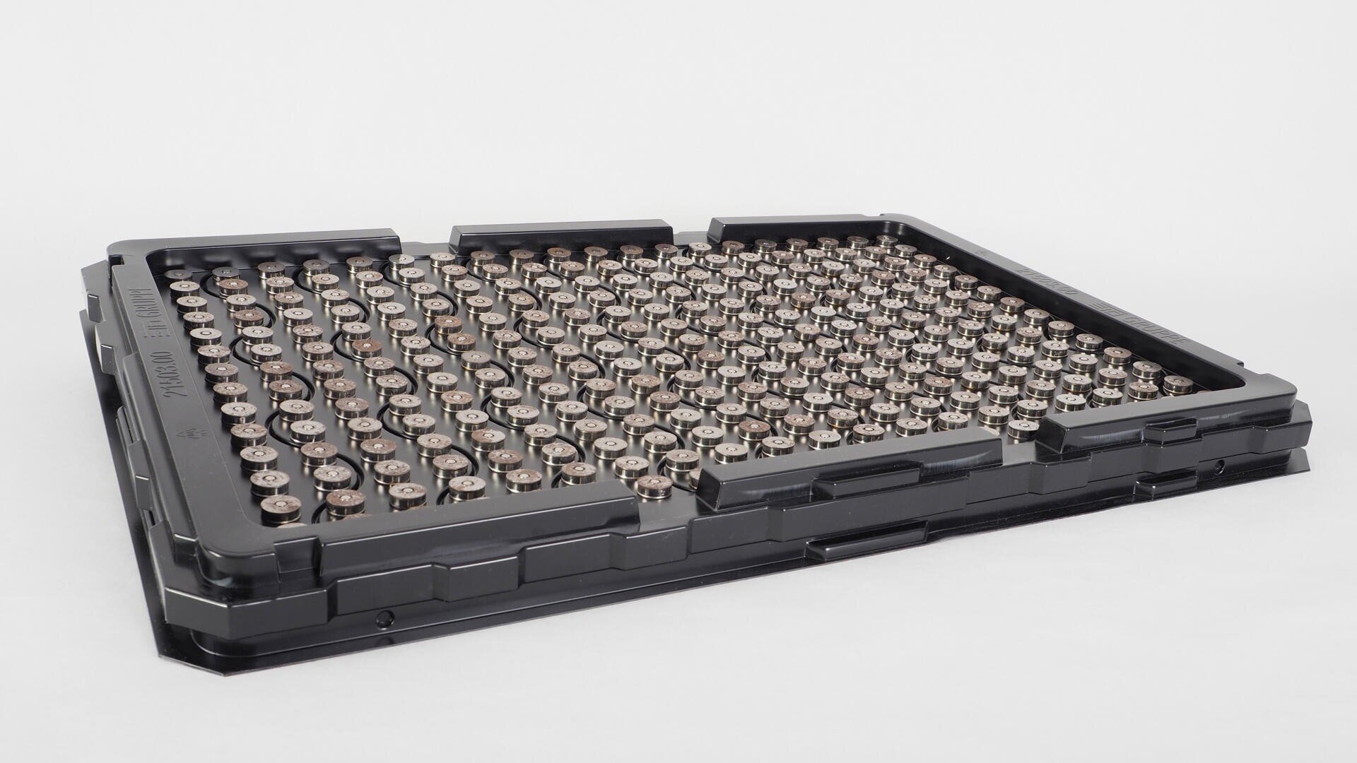 Ein schwarzes Kunststofftablett enthält ein Gitter aus kleinen, zylindrischen Metallobjekten, die in Reihen und Spalten angeordnet sind und schräg auf einem weißen Hintergrund stehen.