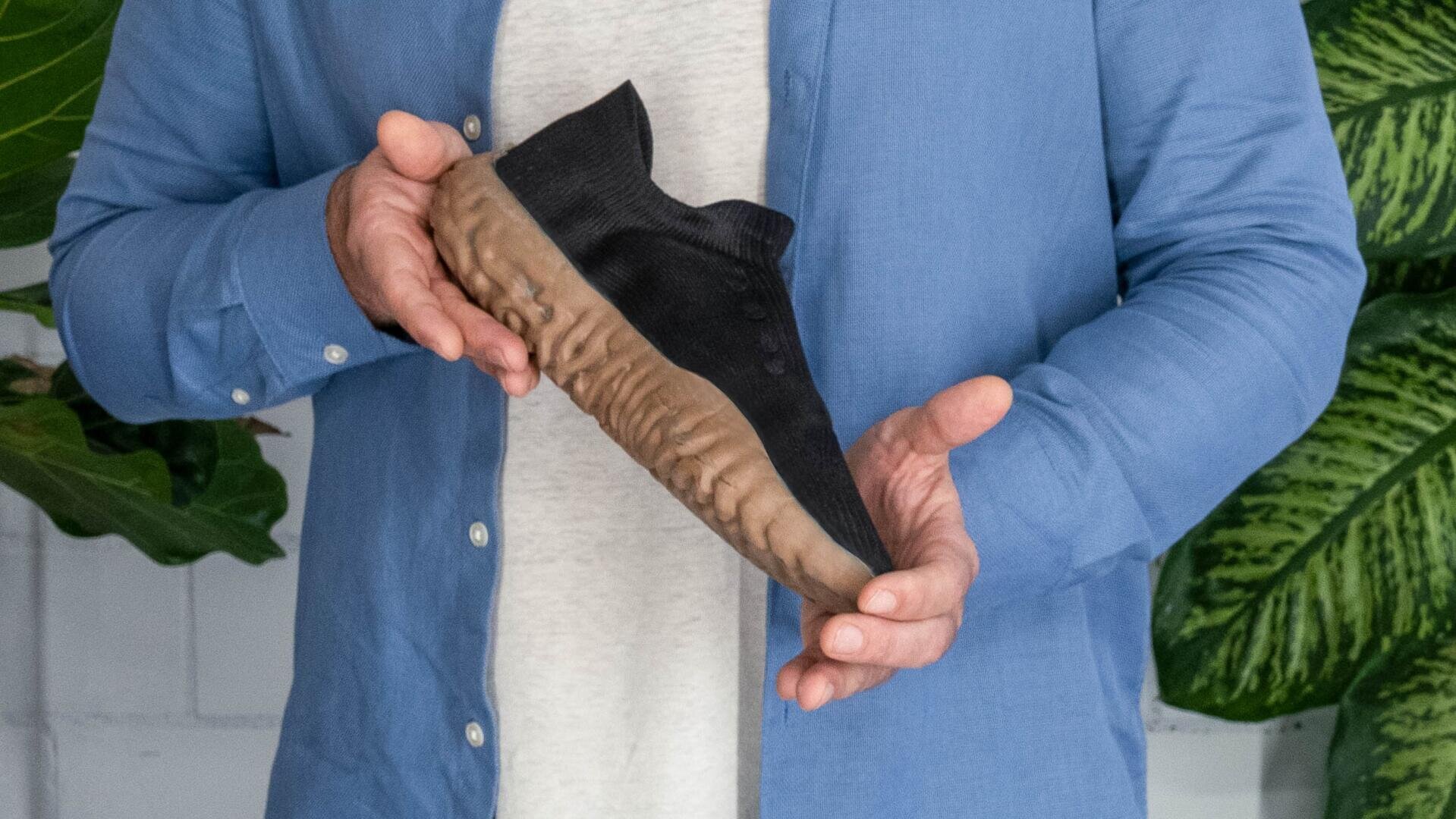 Eine Person in einem blauen Hemd hält einen schwarzen Schuh mit einer dicken, strukturierten braunen Sohle. Im Hintergrund sind grüne Blattpflanzen zu sehen.
