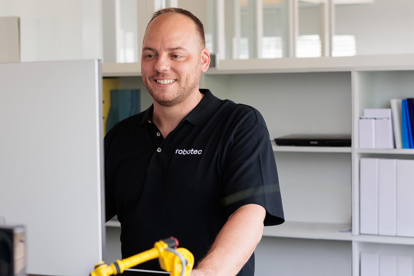 Ein lächelnder Mann in einem schwarzen Robatec-Poloshirt steht an einem Schreibtisch und schaut auf einen Computerbildschirm. Im Hintergrund sind Büroregale und Aktenordner zu sehen.