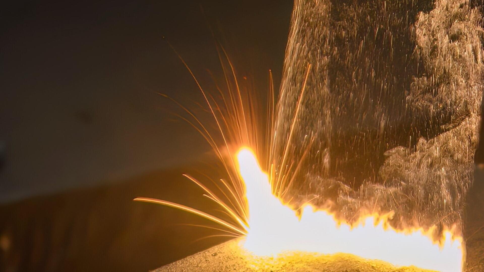 Le métal en fusion se déverse en un flot lumineux et incandescent et des étincelles descendent du point d'impact, créant une scène intense et enflammée sur un fond sombre.