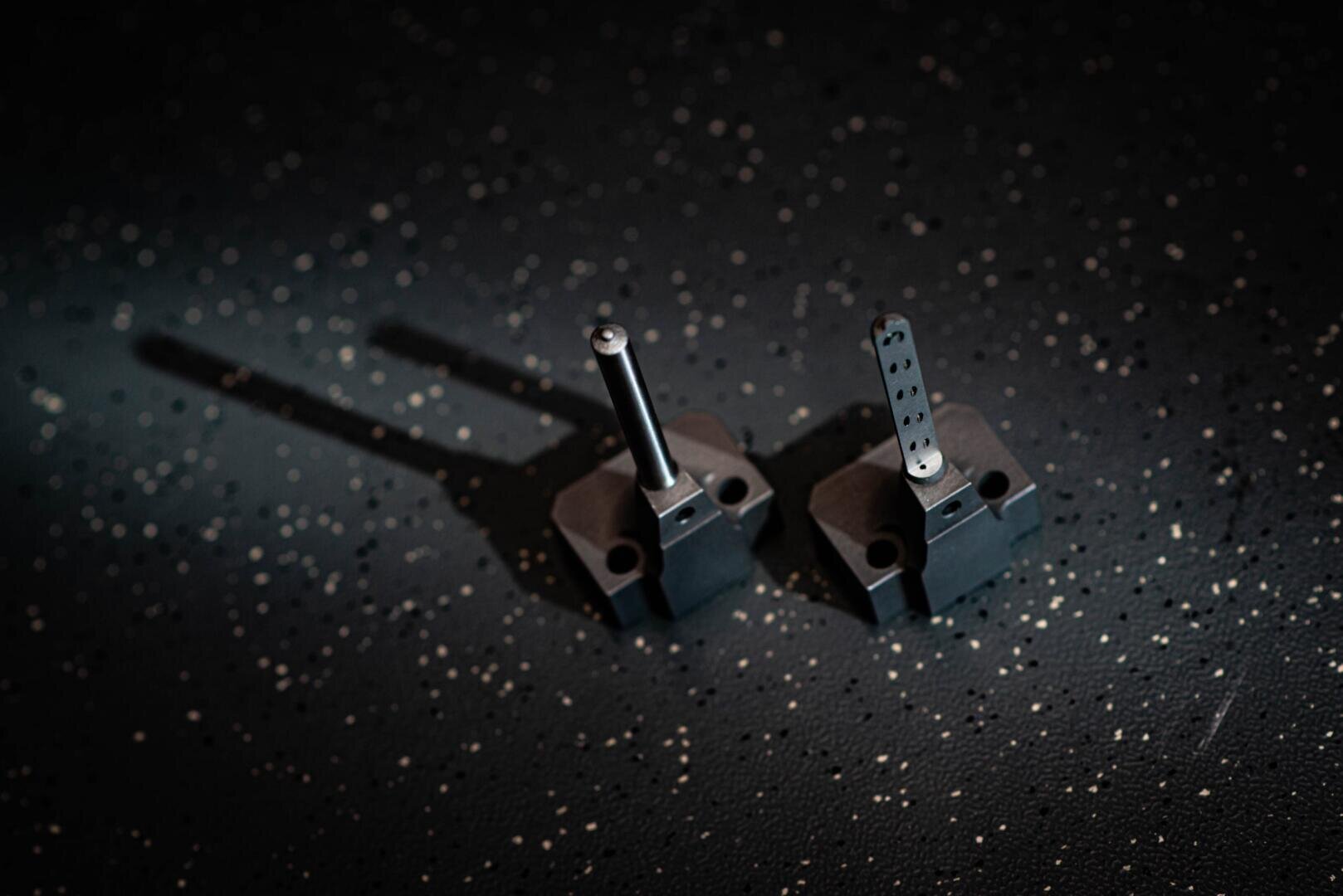 Deux petits composants mécaniques noirs avec des tiges métalliques verticales sont placés sur une surface sombre et mouchetée sous une lumière focalisée qui projette de longues ombres derrière eux.