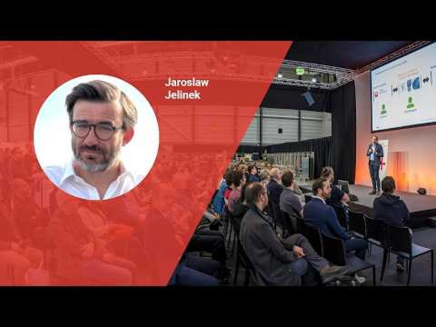 Ein Redner spricht auf der Bühne zu einem Publikum in einem Konferenzraum. Auf der linken Seite zeigt eine rote Einblendung das Porträt eines Mannes mit Brille und dem Namen Jaroslaw Jelinek.
