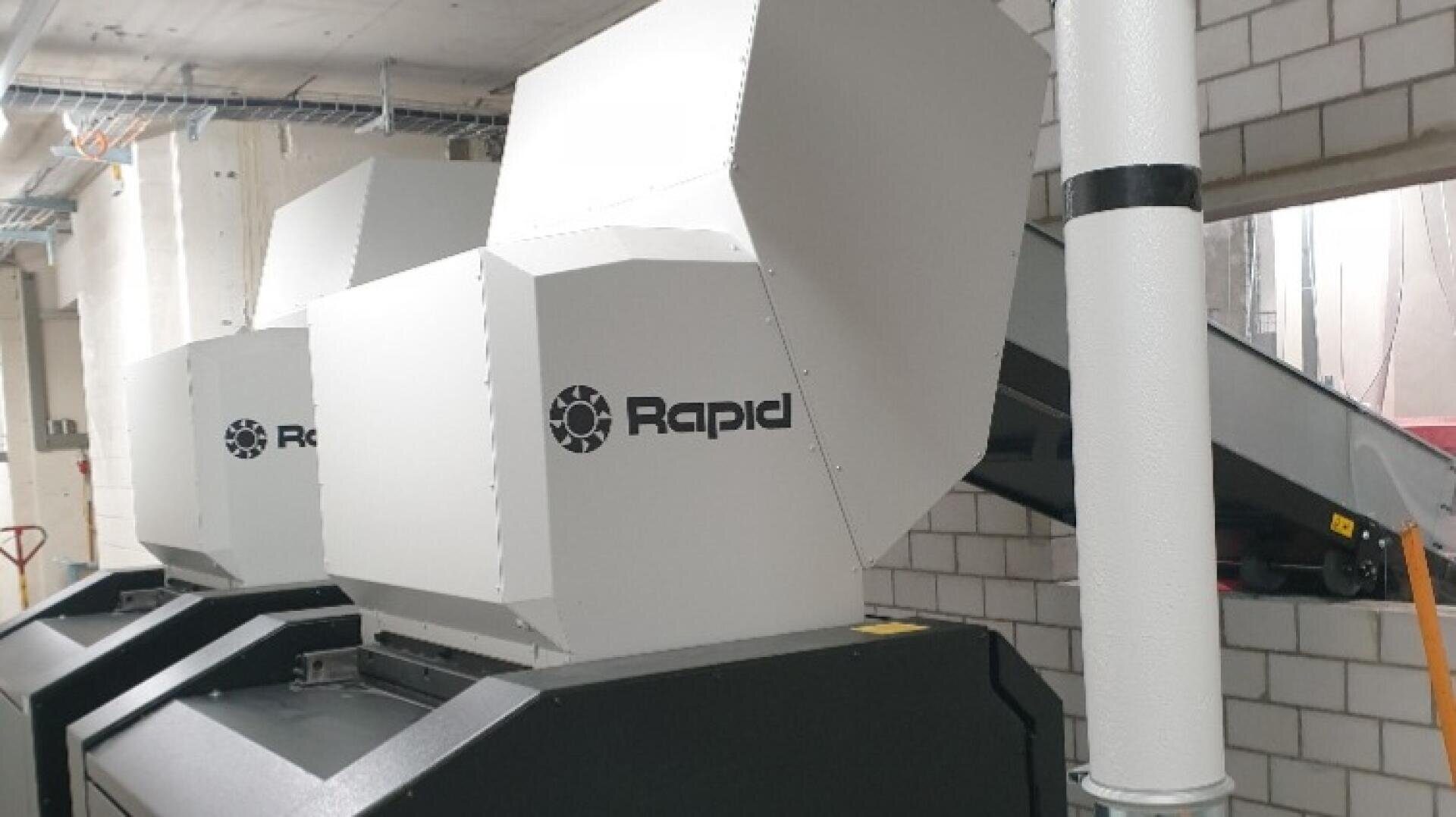 Zwei große Industriemaschinen mit der Aufschrift Rapid sind in einem Innenraum in der Nähe einer weißen Ziegelwand aufgestellt, wobei im Hintergrund ein Teil eines Förderbandes und Rohre zu sehen sind.
