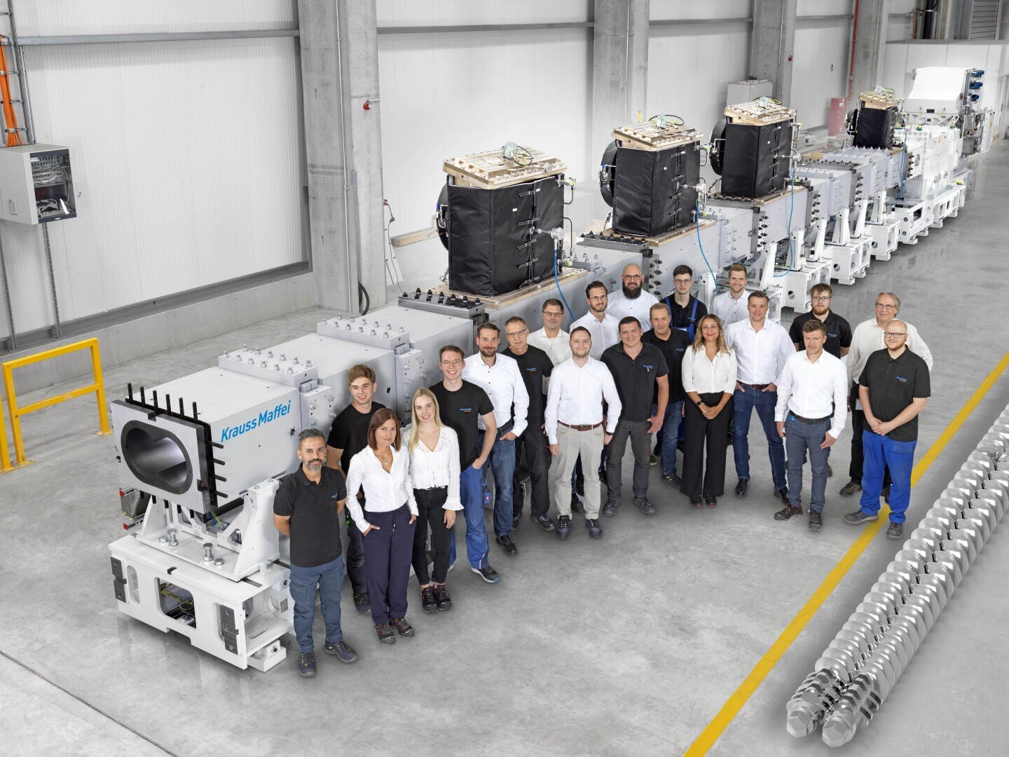 Eine Gruppe von Menschen in Arbeitskleidung steht in einer Fabrikumgebung zusammen und posiert für ein Gruppenfoto vor großen Industriemaschinen und -anlagen. Die Umgebung ist sauber und gut beleuchtet.