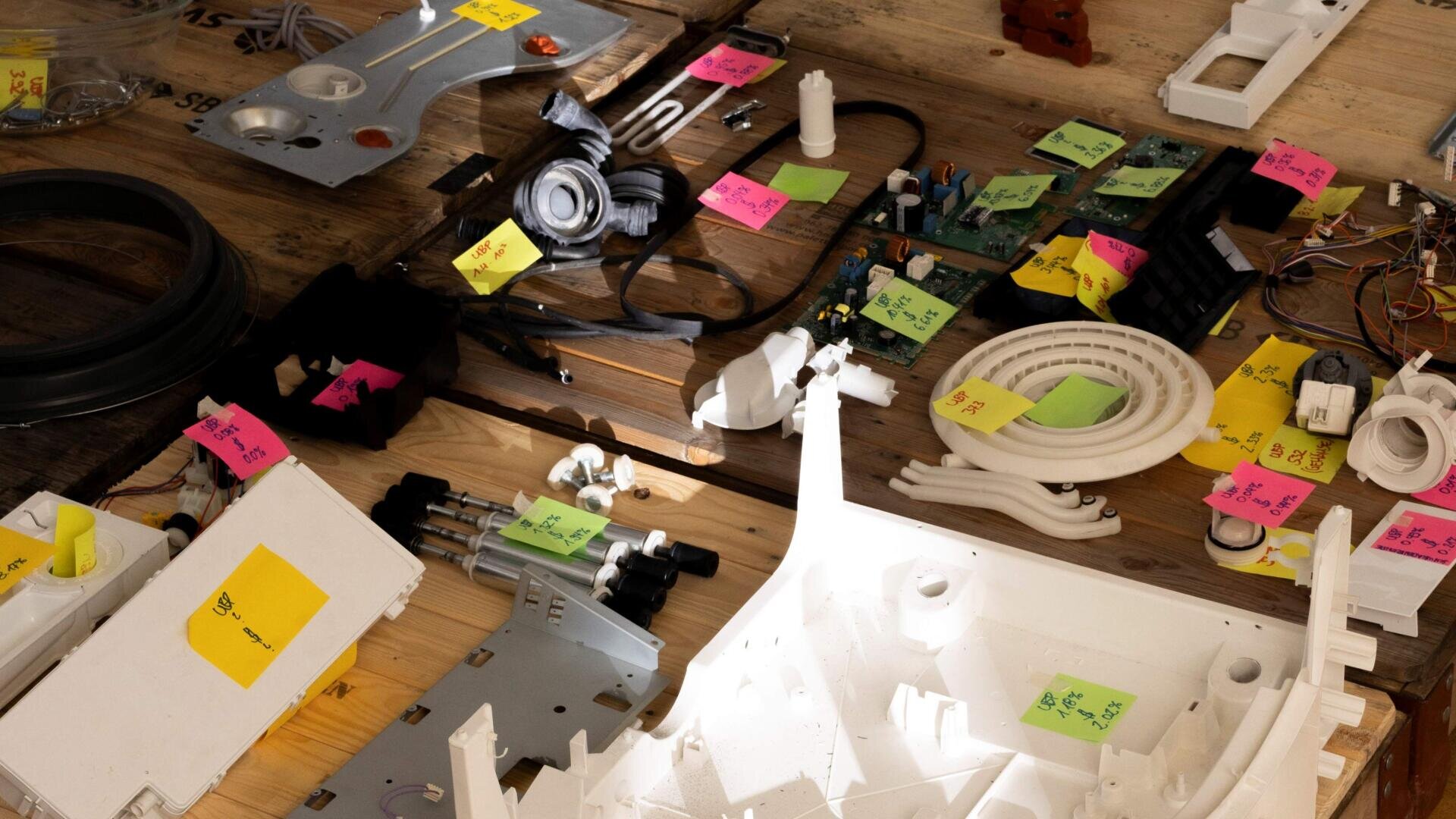 Diverses pièces électroniques et mécaniques sont étalées sur une surface en bois, chacune étiquetée avec des notes autocollantes colorées indiquant leur nom ou leur fonction. Les composants varient en taille et en forme.