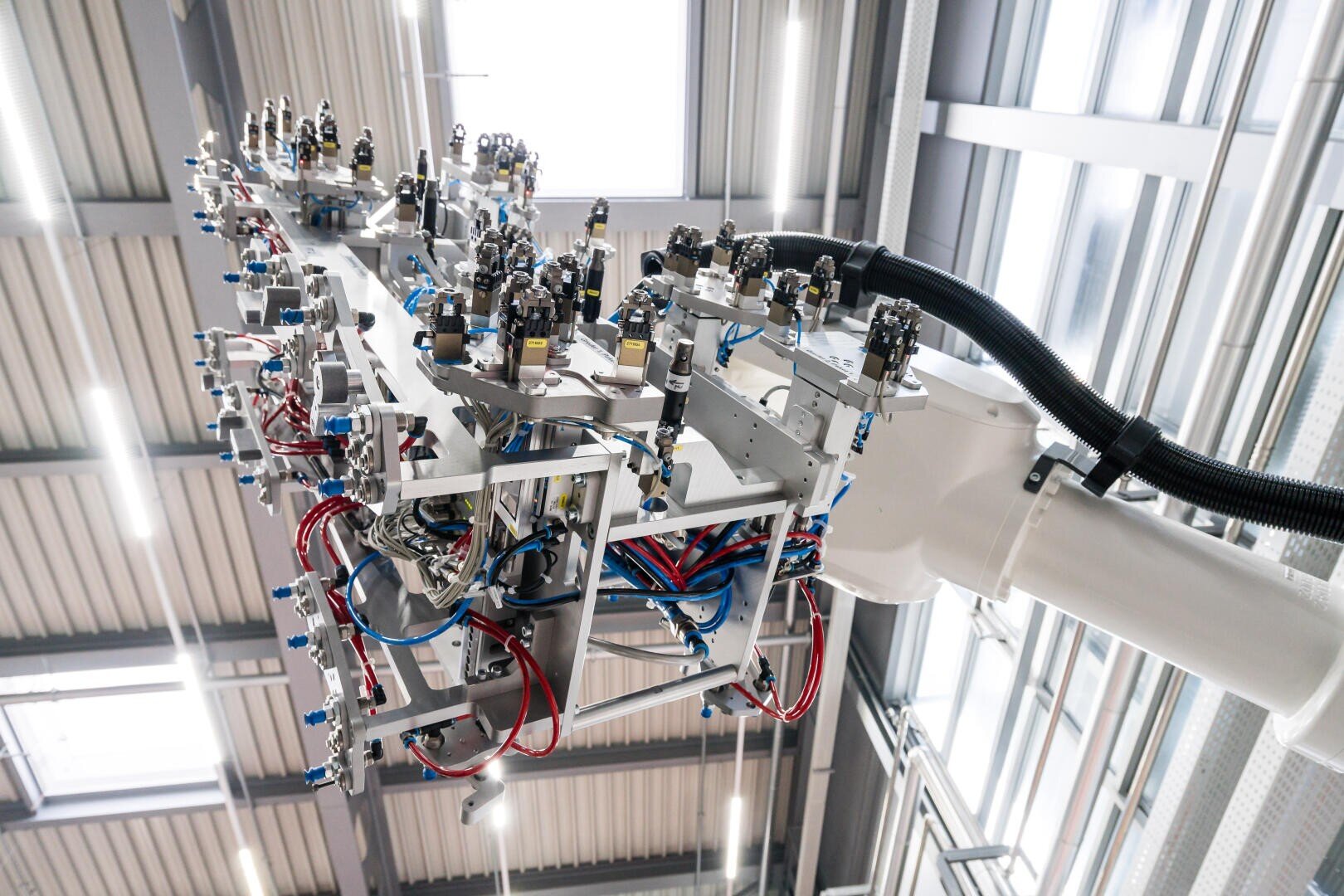 Gros plan sur un bras de robot industriel avec un câblage complexe, des tuyaux et plusieurs capteurs ou composants dans une usine moderne et bien éclairée avec des poutres métalliques et de grandes fenêtres en arrière-plan.