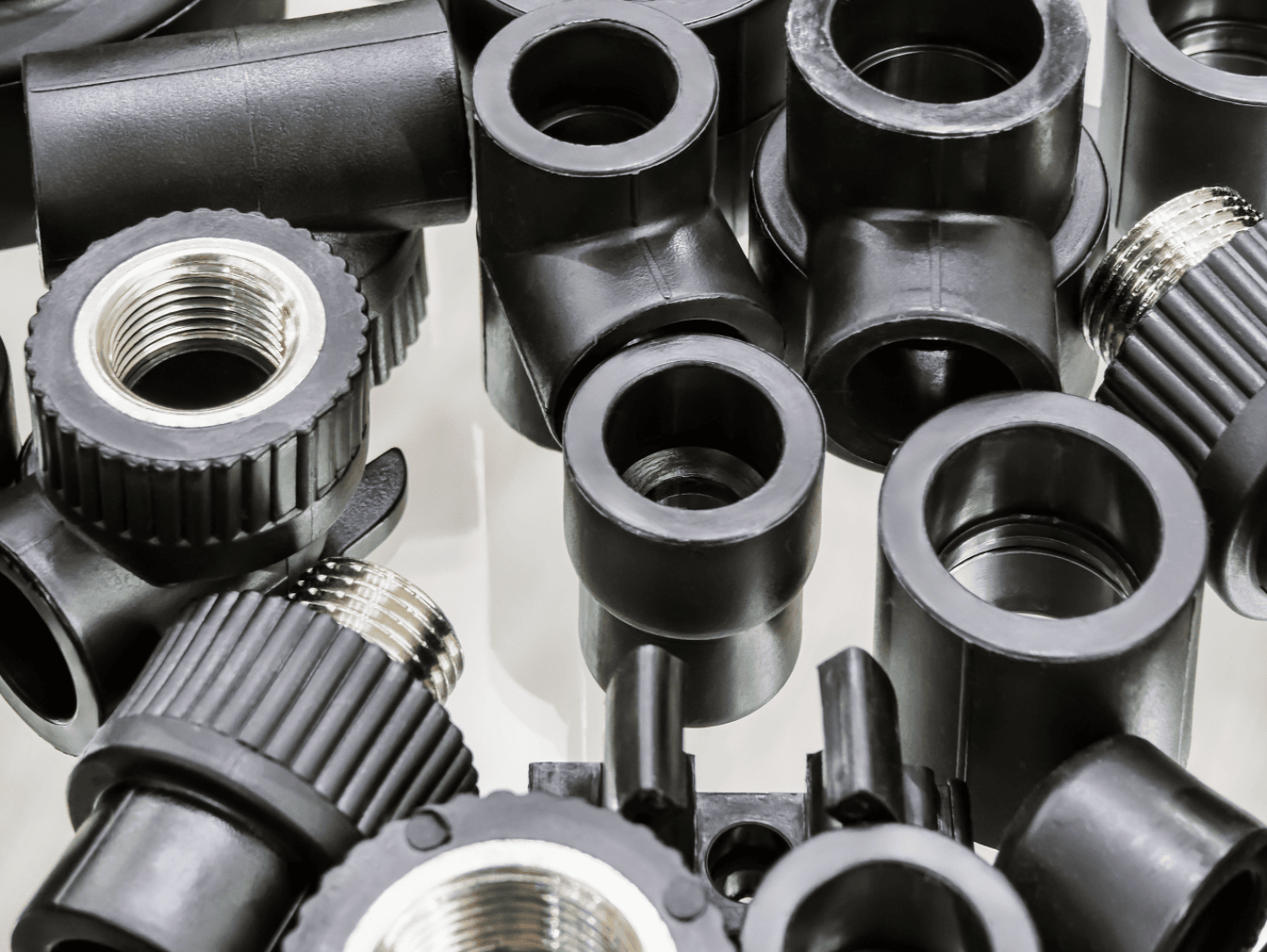 Nahaufnahme mehrerer schwarzer Kunststoffrohrverschraubungen und -verbinder mit Metallgewinde, die in einem zufälligen Stapel auf einer reflektierenden Oberfläche angeordnet sind.