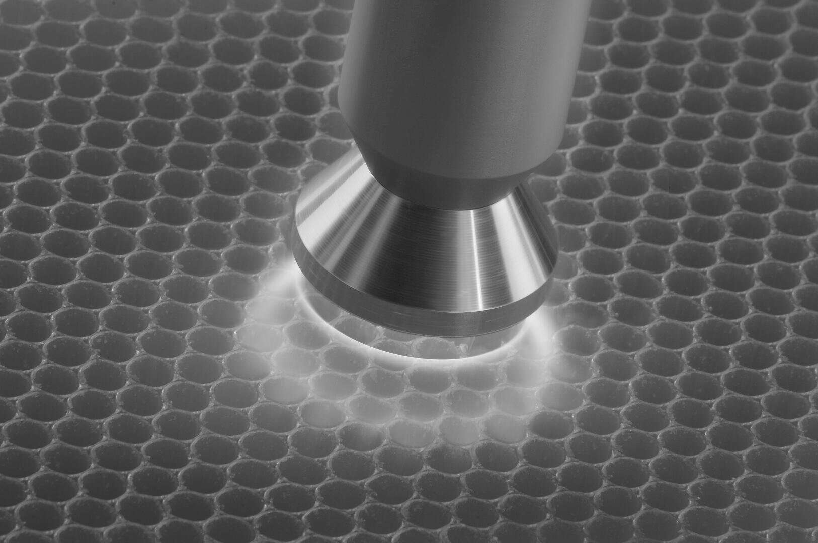 Gros plan sur une buse métallique émettant une lumière blanche et située au-dessus d'une surface en nid d'abeille. La scène a un aspect monochrome très contrasté.