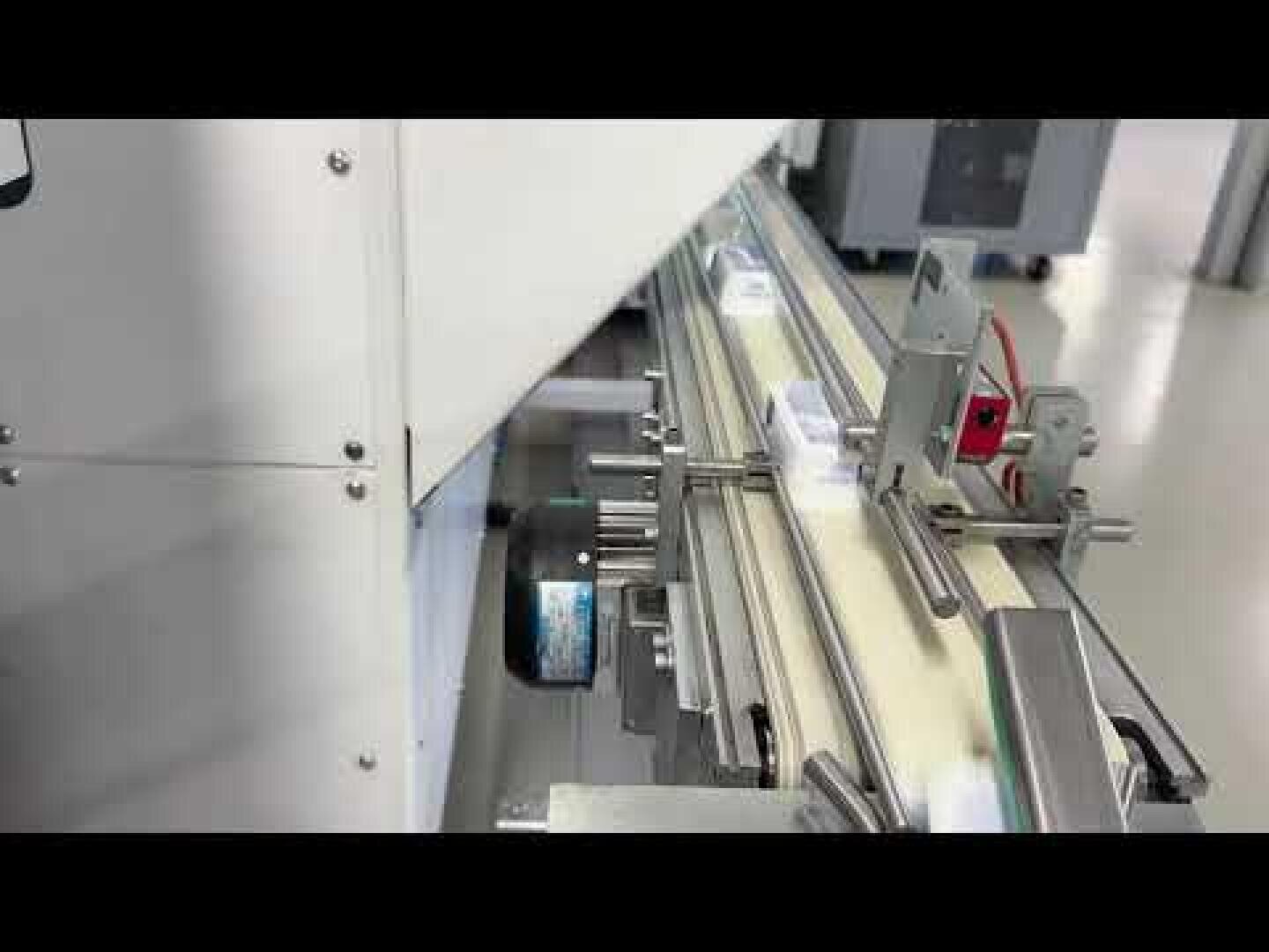 Ein Werksfördersystem mit Metallschienen und -führungen transportiert kleine weiße, rechteckige Produkte durch eine automatische Verarbeitungs- oder Verpackungsmaschine in einer sauberen Industrieumgebung.