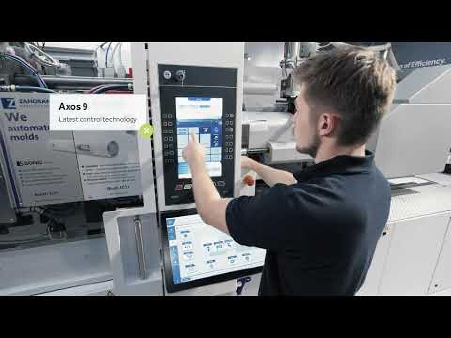 Ein Mann bedient ein modernes Bedienfeld an einer Industriemaschine und interagiert dabei mit einem Touchscreen-Display. Der Bildschirm zeigt den Text "Axos 9 Modernste Steuerungstechnologie" neben technischen Schnittstellenelementen.