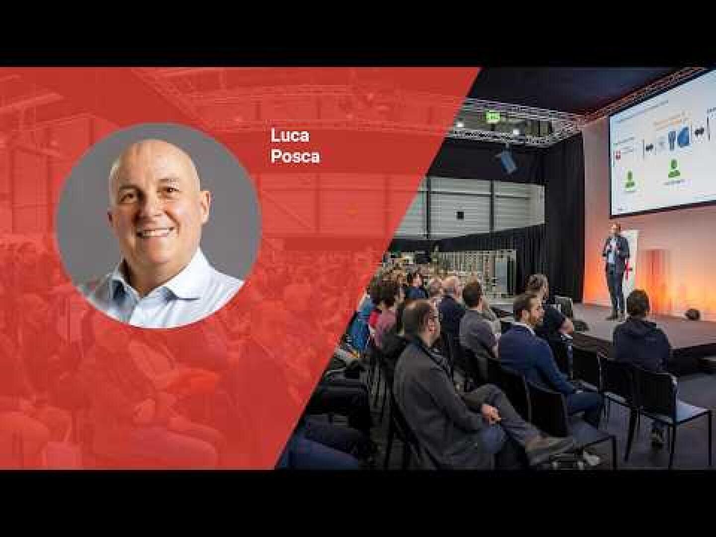 Ein Mann spricht auf der Bühne während einer Konferenz zu einem Publikum. Eine rote Einblendung auf der linken Seite zeigt ein Foto eines lächelnden Mannes, Luca Posca. Hinter dem Redner werden Dias projiziert.