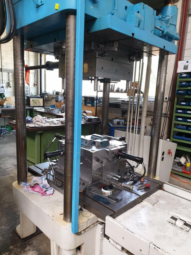 Une grande presse industrielle hydraulique avec un moule métallique sur son banc dans un atelier où l'on peut voir des outils, des étagères et des appareils en arrière-plan.