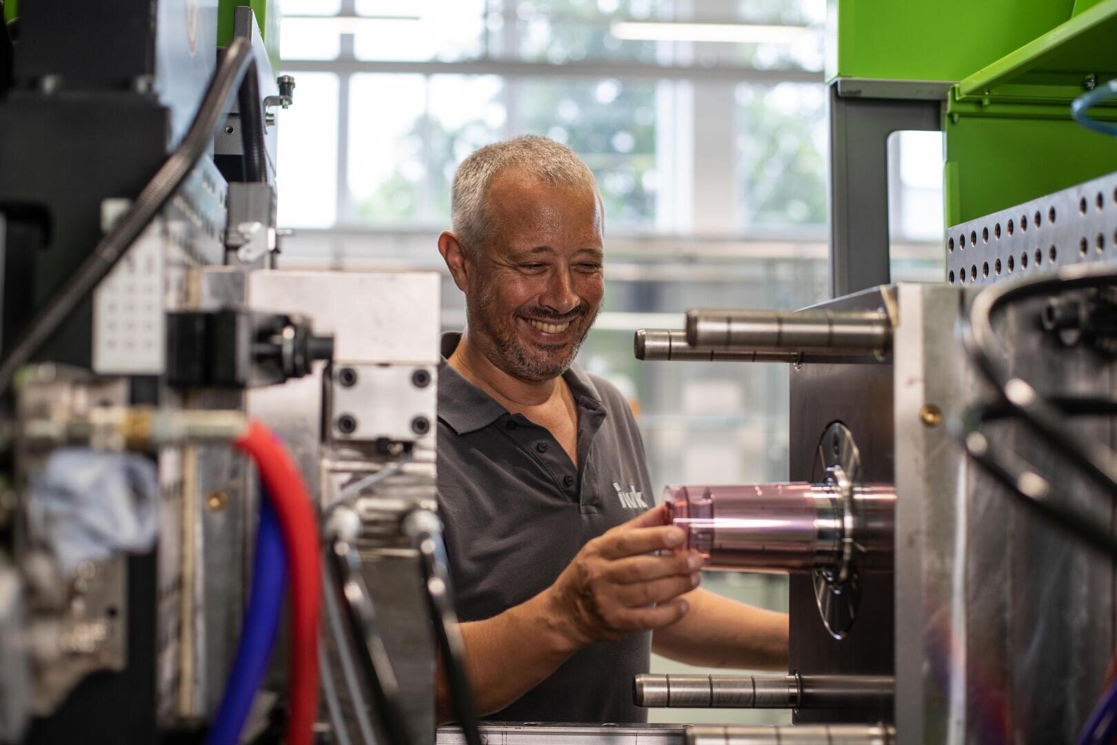 Ein lächelnder Mann bedient Industriemaschinen und platziert einen zylindrischen Gegenstand in einer großen Metallvorrichtung in einer hell erleuchteten Werkstatt mit grünen Akzenten und großen Fenstern im Hintergrund.
