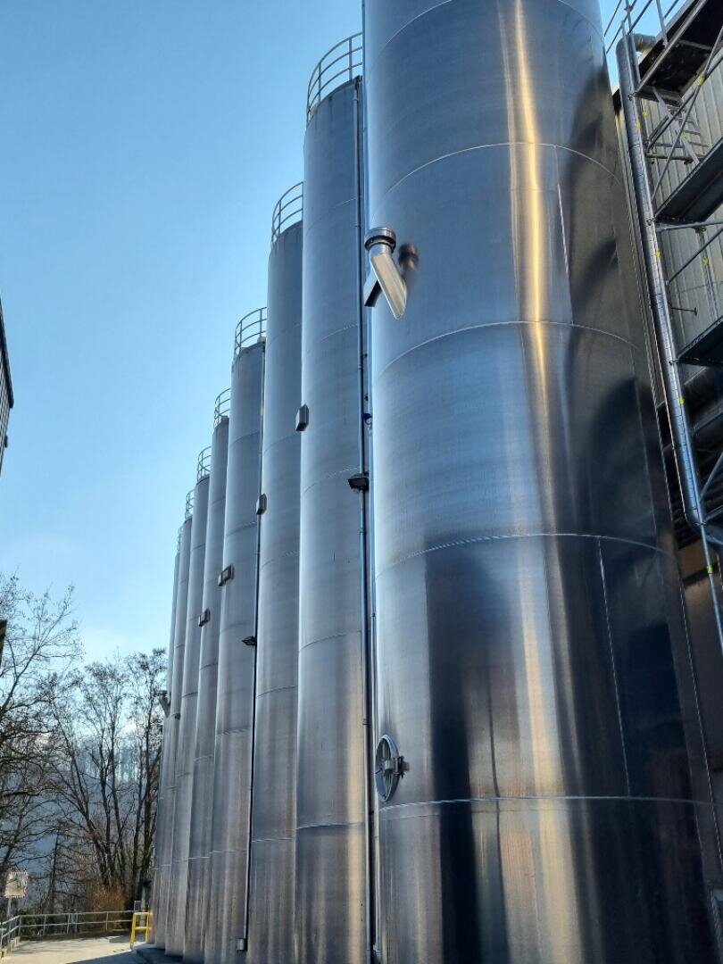 Hohe, glänzende Metalltanks reihen sich im Freien vor einem strahlend blauen Himmel auf, mit einem Geländer obenauf und einigen Bäumen auf der linken Seite.