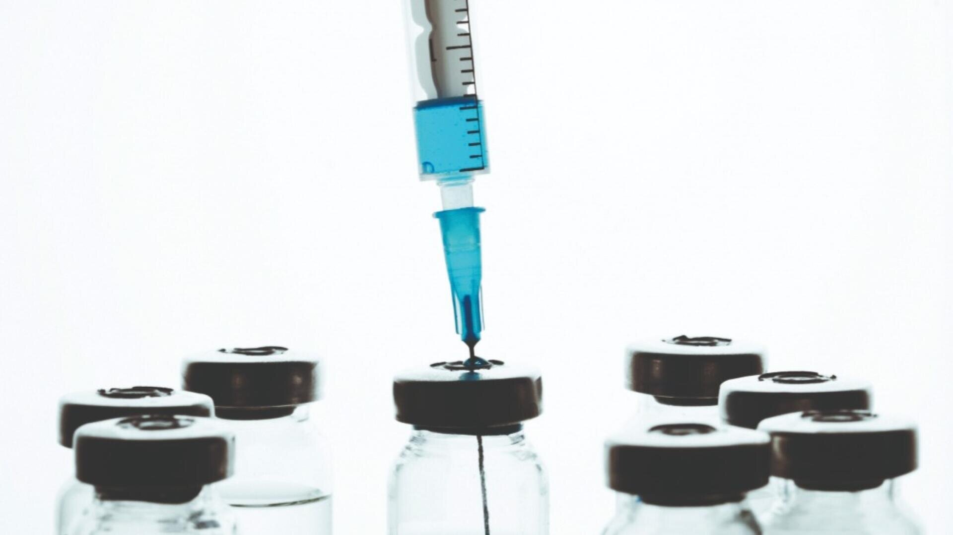 Nahaufnahme einer Spritze, die Flüssigkeit aus einem Fläschchen aufzieht, umgeben von mehreren ähnlichen Fläschchen mit grauen Deckeln, vor einem weißen Hintergrund.