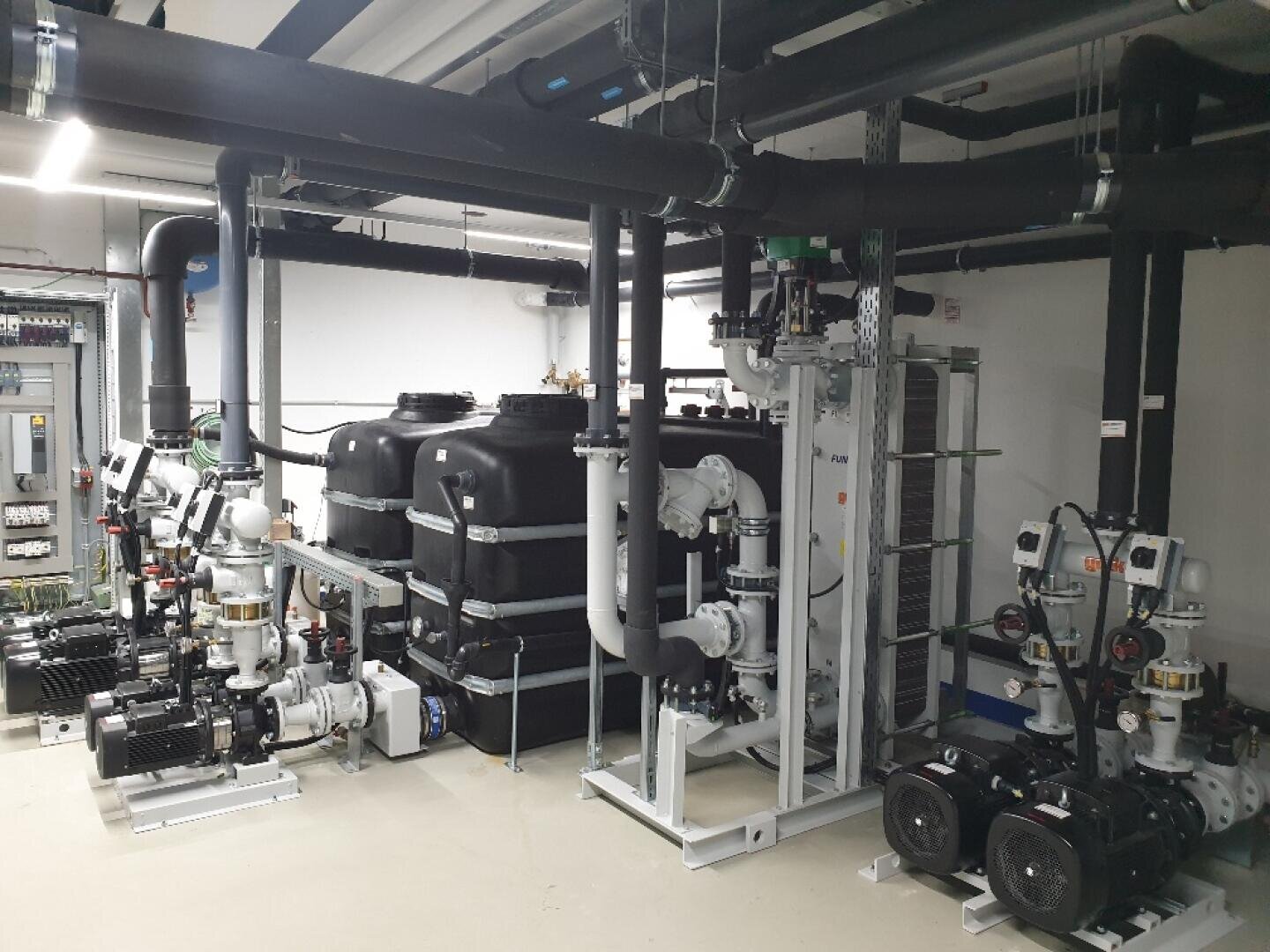 Ein Raum mit industrieller Ausrüstung, darunter große schwarze Tanks, zahlreiche Rohre, Pumpen und Schalttafeln, die ordentlich in einer sauberen, gut beleuchteten Umgebung angeordnet sind.