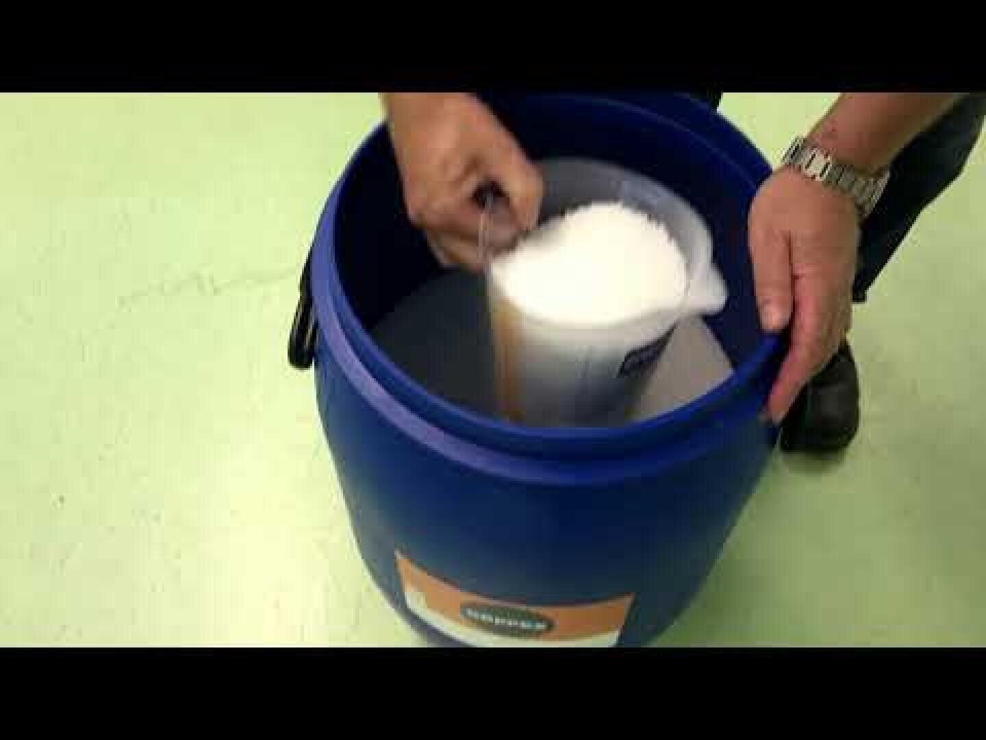 Eine Person schöpft mit einem weißen Messbecher weißes körniges Material, möglicherweise Salz oder eine ähnliche Substanz, aus einem großen blauen Plastikbehälter.