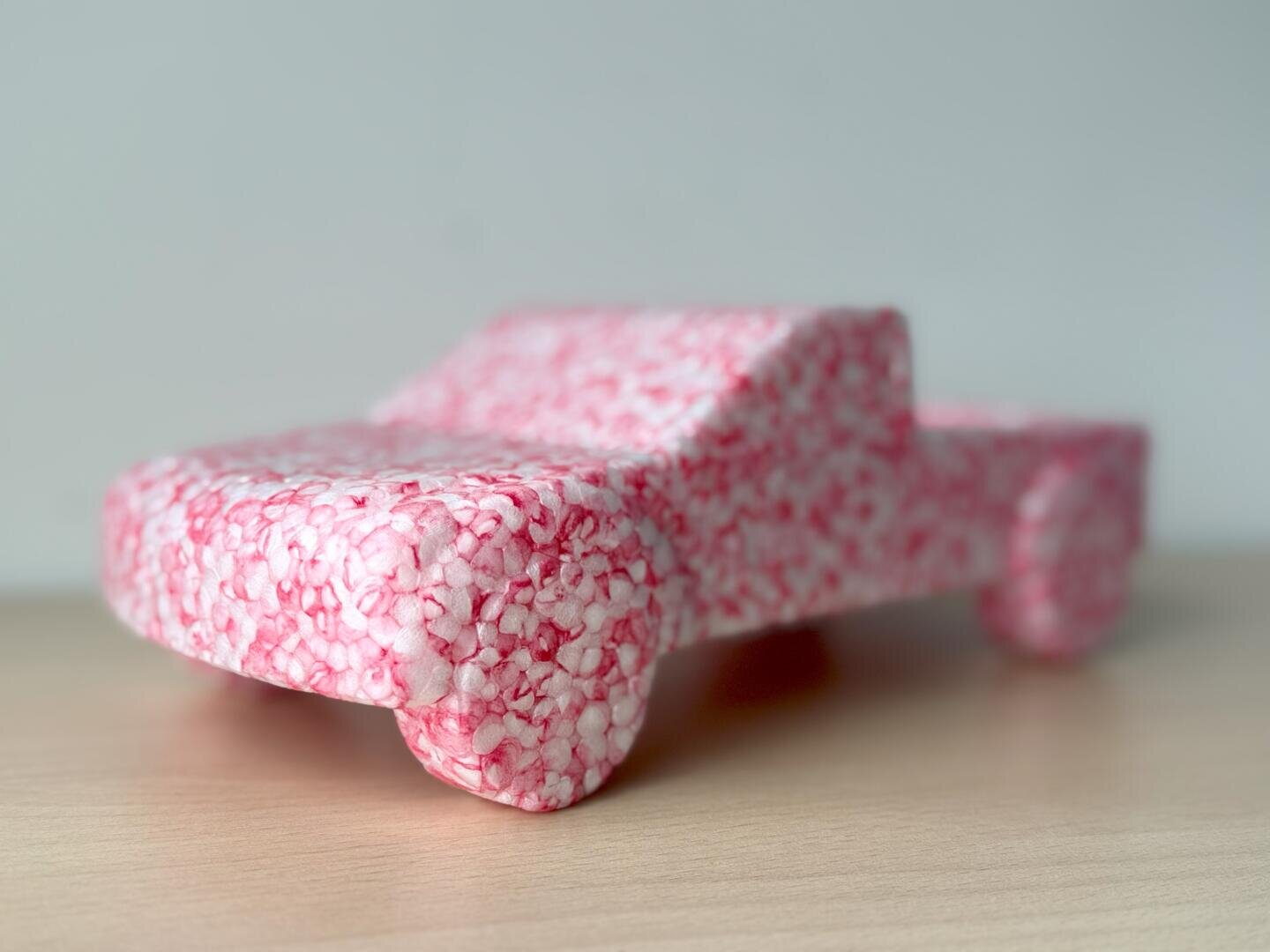 Un camion-jouet en mousse rose et blanche mouchetée est posé sur une surface en bois clair sur un fond sobre gris clair.