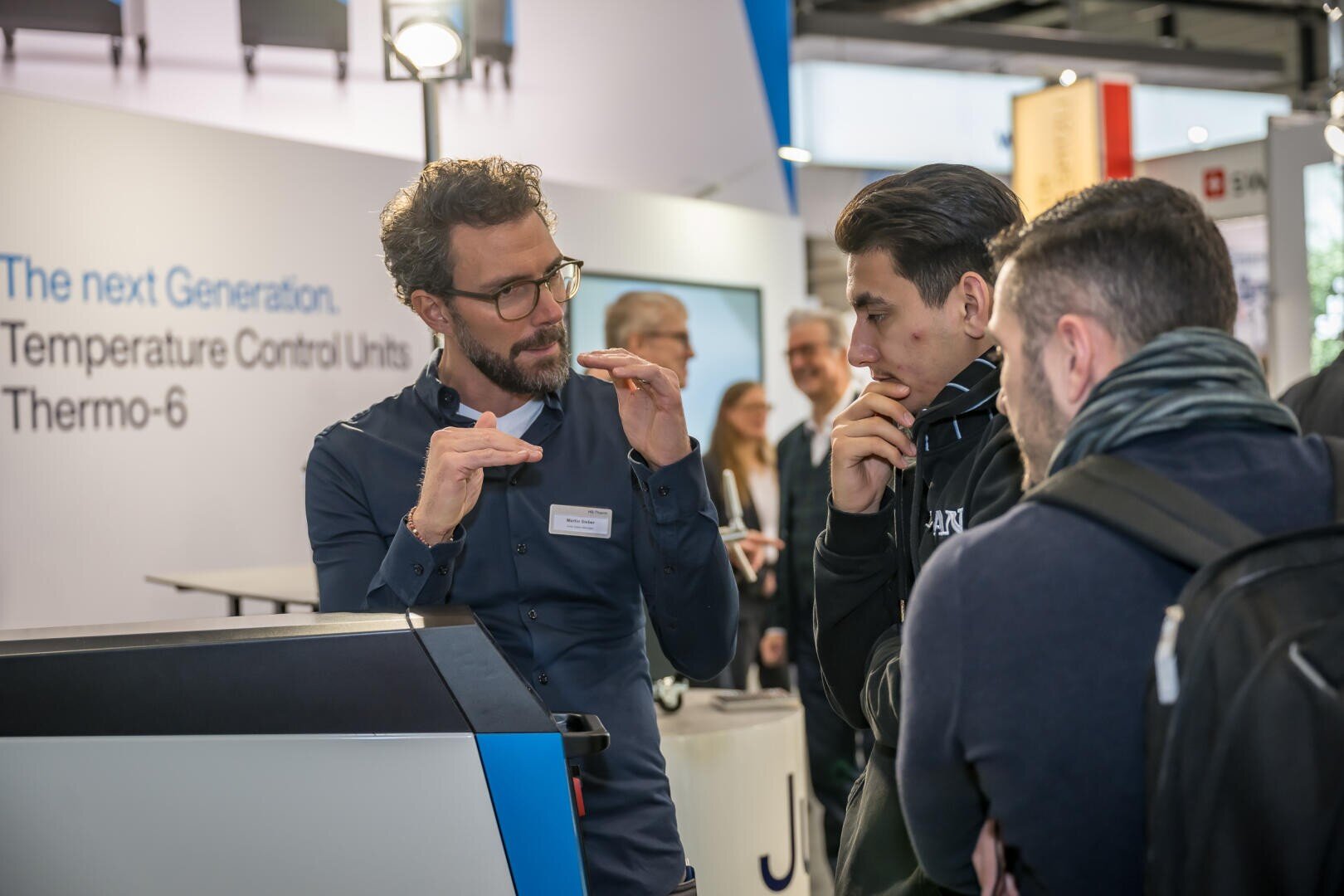 Un homme portant des lunettes explique quelque chose à deux hommes attentifs à un stand technologique à l'aide de gestes de la main. Sur un écran en arrière-plan, on peut lire : La prochaine génération : les thermorégulateurs Thermo-6.