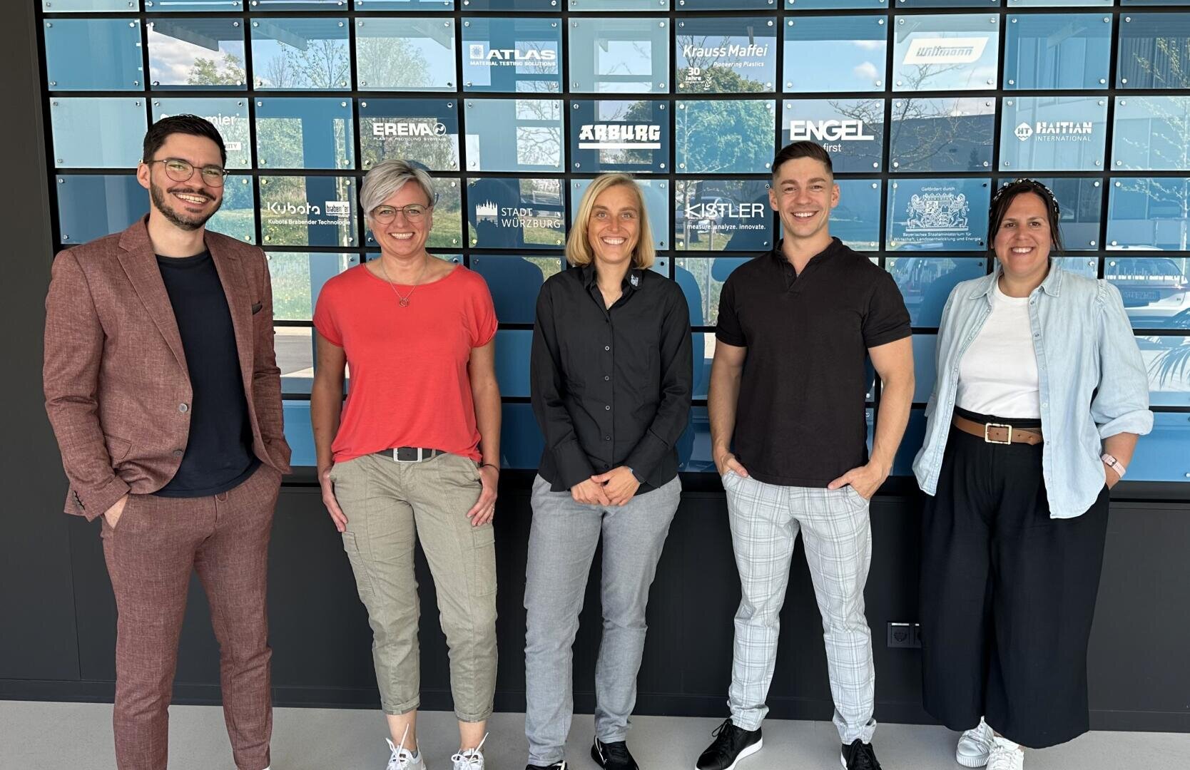 Fünf Personen stehen lächelnd vor einer Wand mit Firmenlogos, gekleidet in legerer Business-Kleidung. Drei Frauen und zwei Männer posieren zusammen in einem hellen, modernen Innenraum.