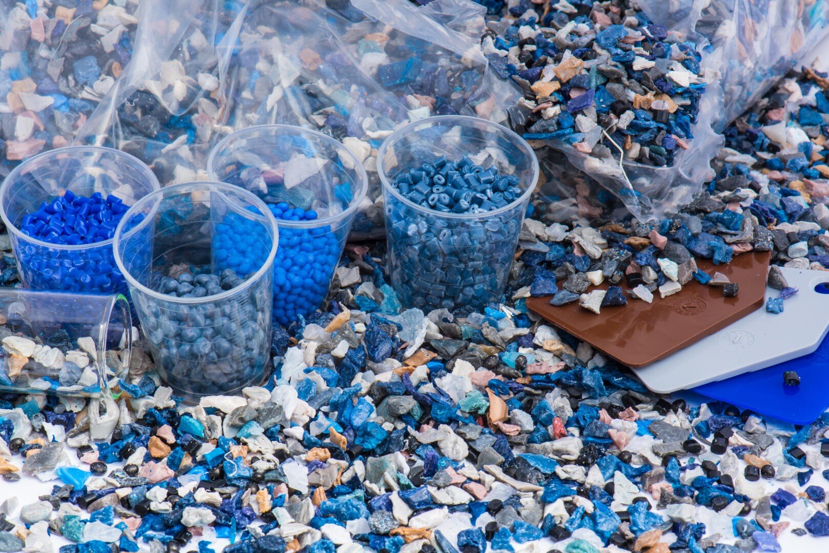 Mehrere durchsichtige Plastikbecher, die mit blauen Plastikkügelchen gefüllt sind, stehen auf einer Fläche, die mit blauen, weißen und braunen Plastikfragmenten bedeckt ist, mit mehreren Farbmusterkarten in der Nähe und Plastiktüten im Hintergrund.