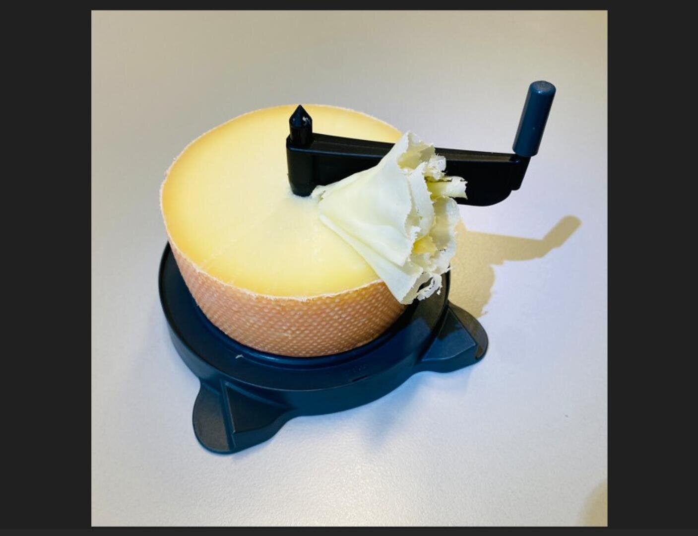 Ein runder Käselaib auf einem schwarzen Lockenwickler mit einem Griff und einer Klinge, die eine dünne, gekräuselte Käselocke auf der Oberseite erzeugt, auf einer hellen Oberfläche platziert.