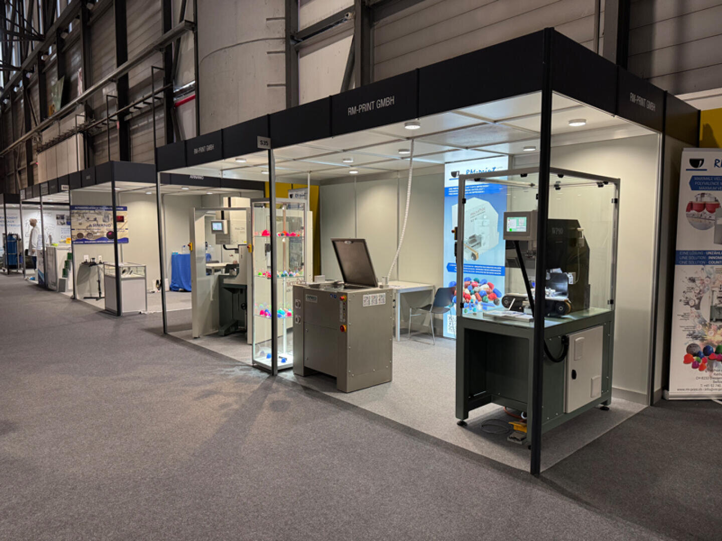 Auf einem Messestand sind verschiedene Industriemaschinen in Glasgehäusen ausgestellt, und es werden Bildschirme und 3D-gedruckte Objekte gezeigt. Der Bereich ist gut beleuchtet, und die benachbarten Stände sind im Hintergrund zu sehen.