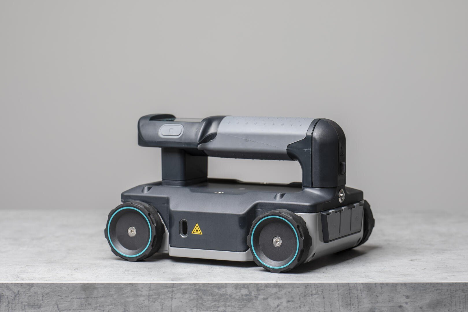Ein kleines, schwarzes Roboterfahrzeug mit vier blau umrandeten Rädern und einem Griff an der Oberseite steht auf einer hellgrauen Fläche vor einem schlichten, hellgrauen Hintergrund.