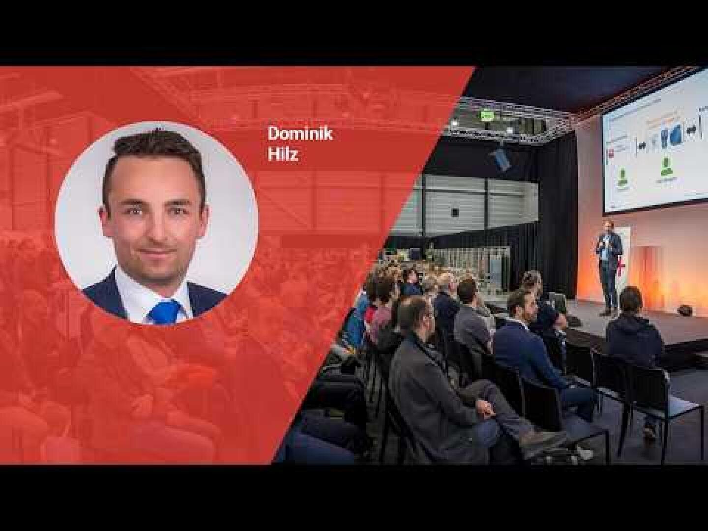 Un conférencier s'adresse à un public sur scène. Sur la gauche, une incrustation rouge montre la photo d'un homme en costume, Dominik Hilz. La salle est composée de rangées de participants assis et d'écrans géants.
