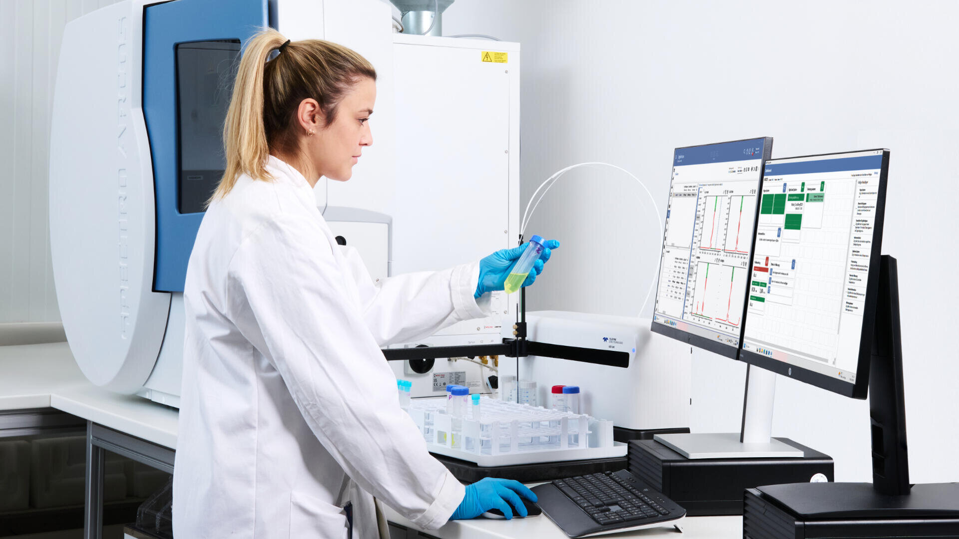 Ein Wissenschaftler in einem weißen Laborkittel und mit blauen Handschuhen analysiert ein Reagenzglas an einem Arbeitsplatz, der von Laborgeräten umgeben ist und dessen Daten auf zwei Computermonitoren angezeigt werden.