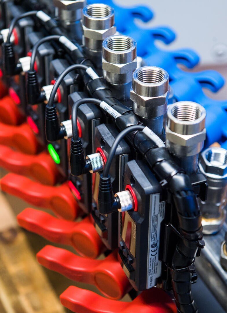 Nahaufnahme einer Reihe von Metallventilen und -anschlüssen mit roten Griffen und blauen Kunststoffteilen, an denen schwarze Drähte und elektronische Komponenten befestigt sind.