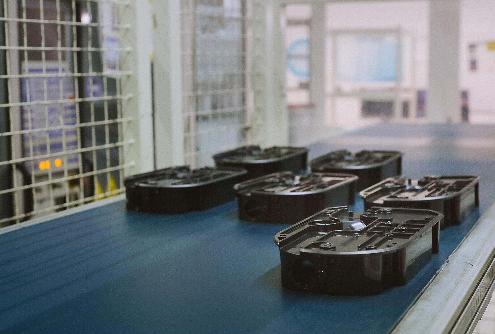 Sechs schwarze Kunststoffteile sind auf einem blauen Förderband in einer Fabrik oder Produktionsstätte aufgereiht, im Hintergrund sind weiße Metallschutzwände und Industrieanlagen zu sehen.