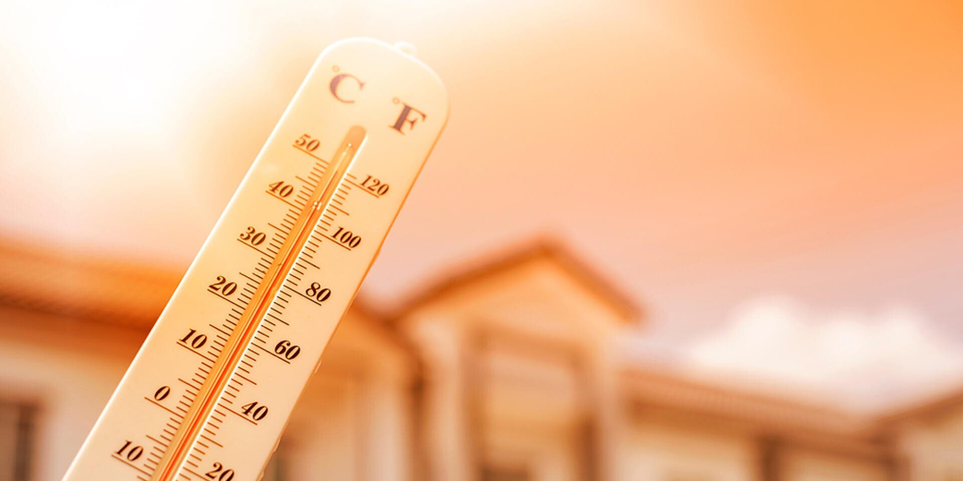 Un thermomètre indiquant une température élevée, sur un fond flou avec une maison et un ciel orange vif, indiquant une journée chaude et ensoleillée.