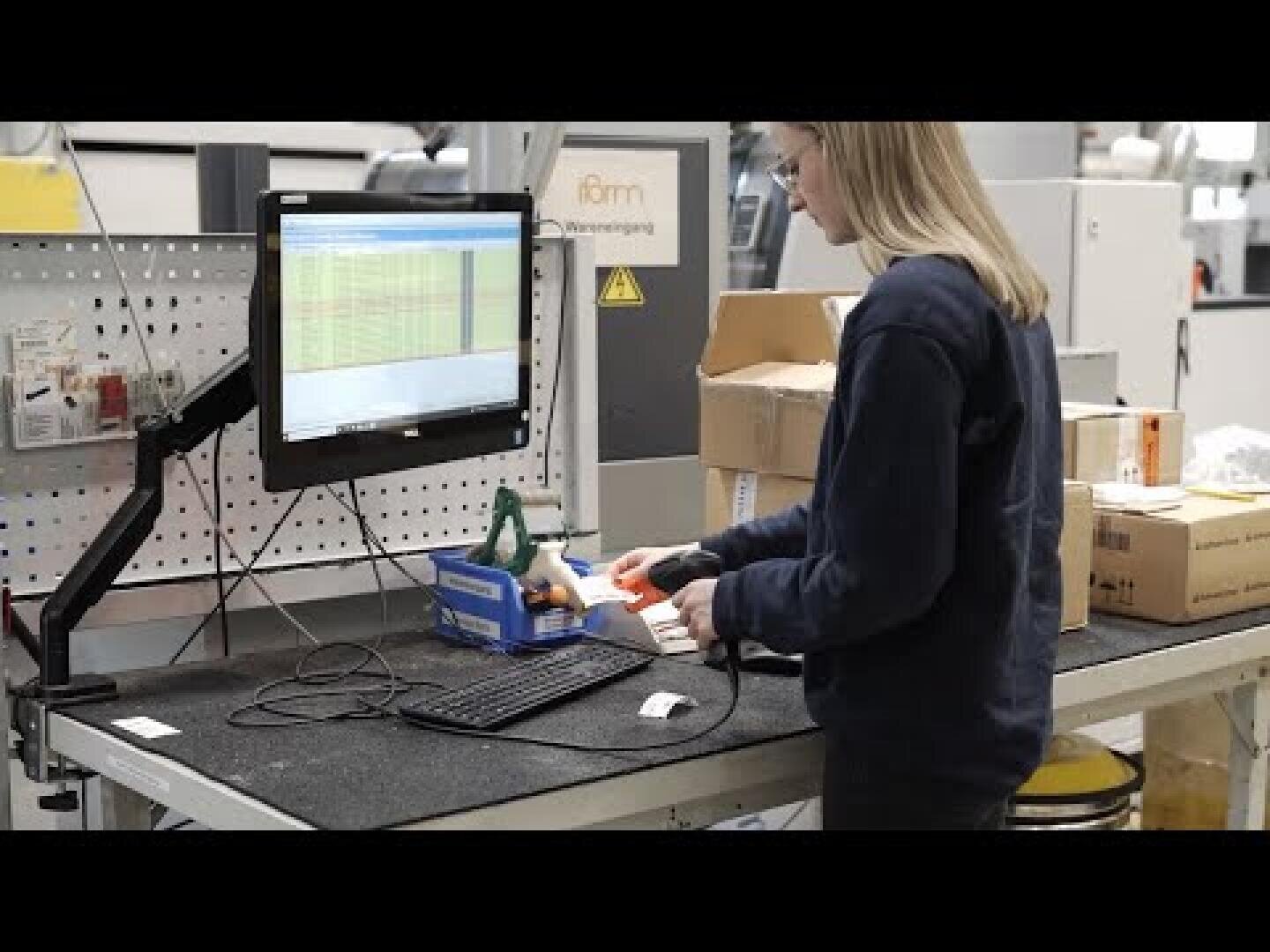 Eine Person benutzt einen Barcode-Scanner an einem Arbeitsplatz mit einem Computermonitor, der Daten anzeigt, umgeben von Kartons und Büromaterial in einer industriellen Umgebung.