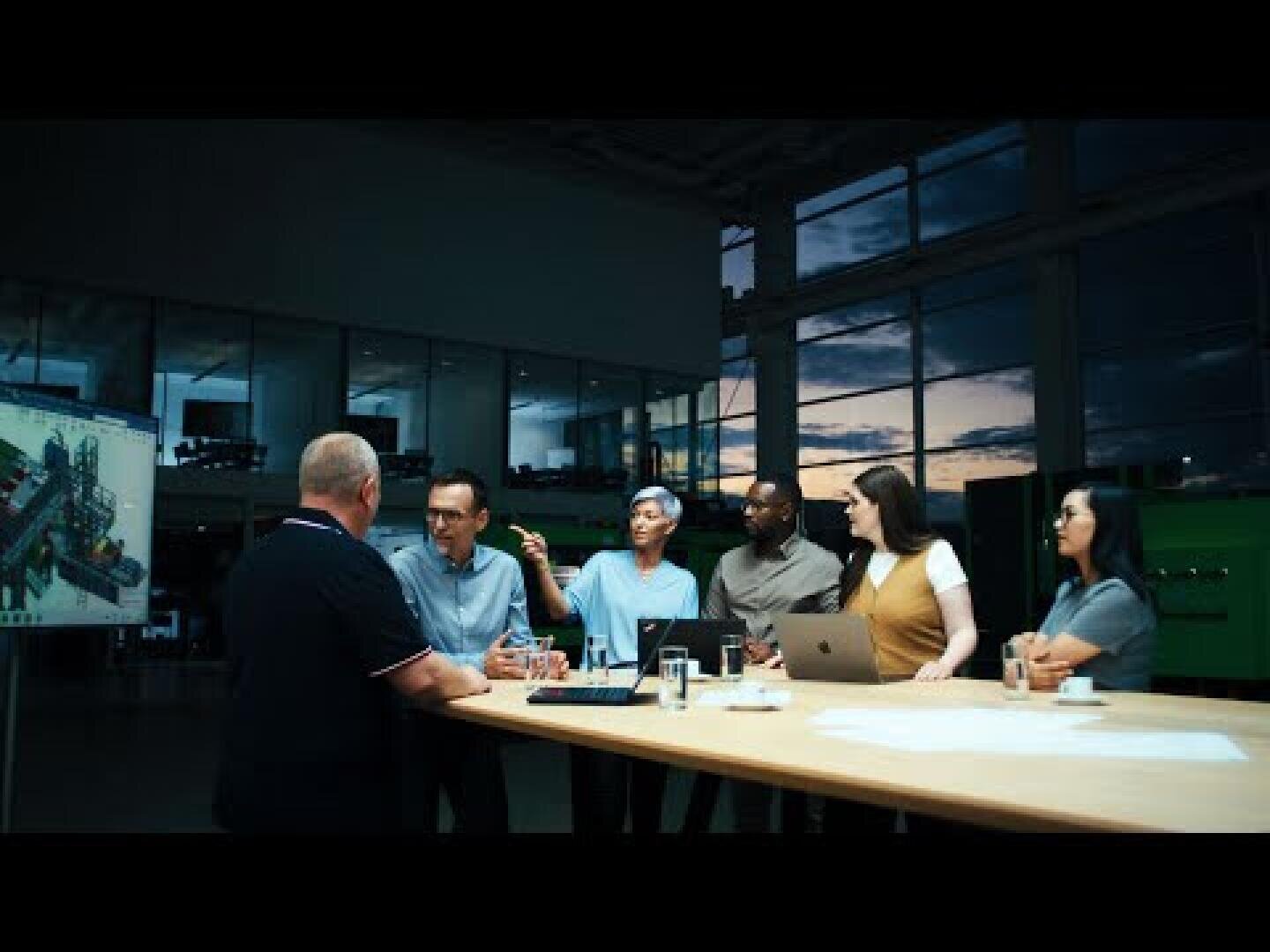 Eine Gruppe von sechs Personen sitzt und steht in der Abenddämmerung um einen großen Tisch in einem modernen Büro und diskutiert. Auf dem Tisch befinden sich Laptops, Dokumente und Getränke. Eine Blaupause oder ein Bild wird auf eine Leinwand im Hintergrund projiziert.