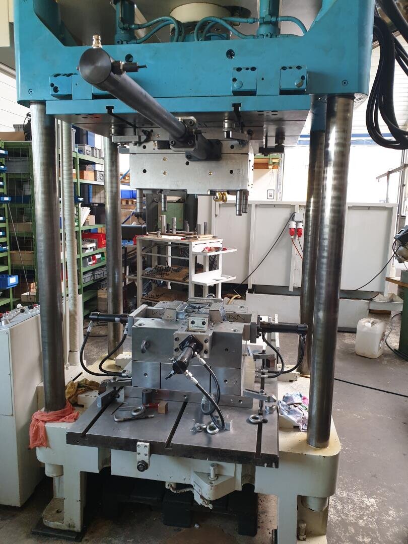 Une grande presse hydraulique industrielle avec des pièces métalliques et des tuyaux dans un atelier, entourée d'outils, d'étagères et d'équipements. La machine est posée sur une plateforme métallique et est prête à fonctionner.