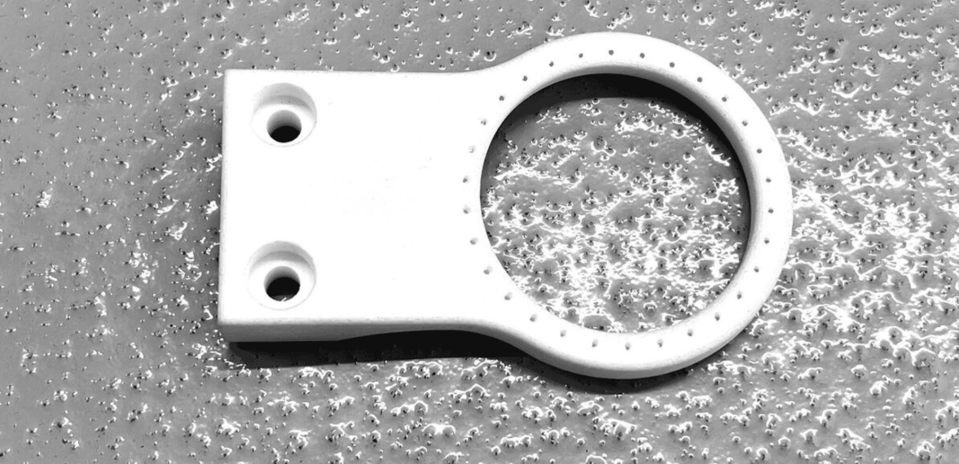 Une anse blanche, plate et circulaire, avec deux trous de vis sur un côté, repose sur une surface texturée et mouchetée de gris. L'anse a une forme annulaire avec de petites bosses sur le bord.