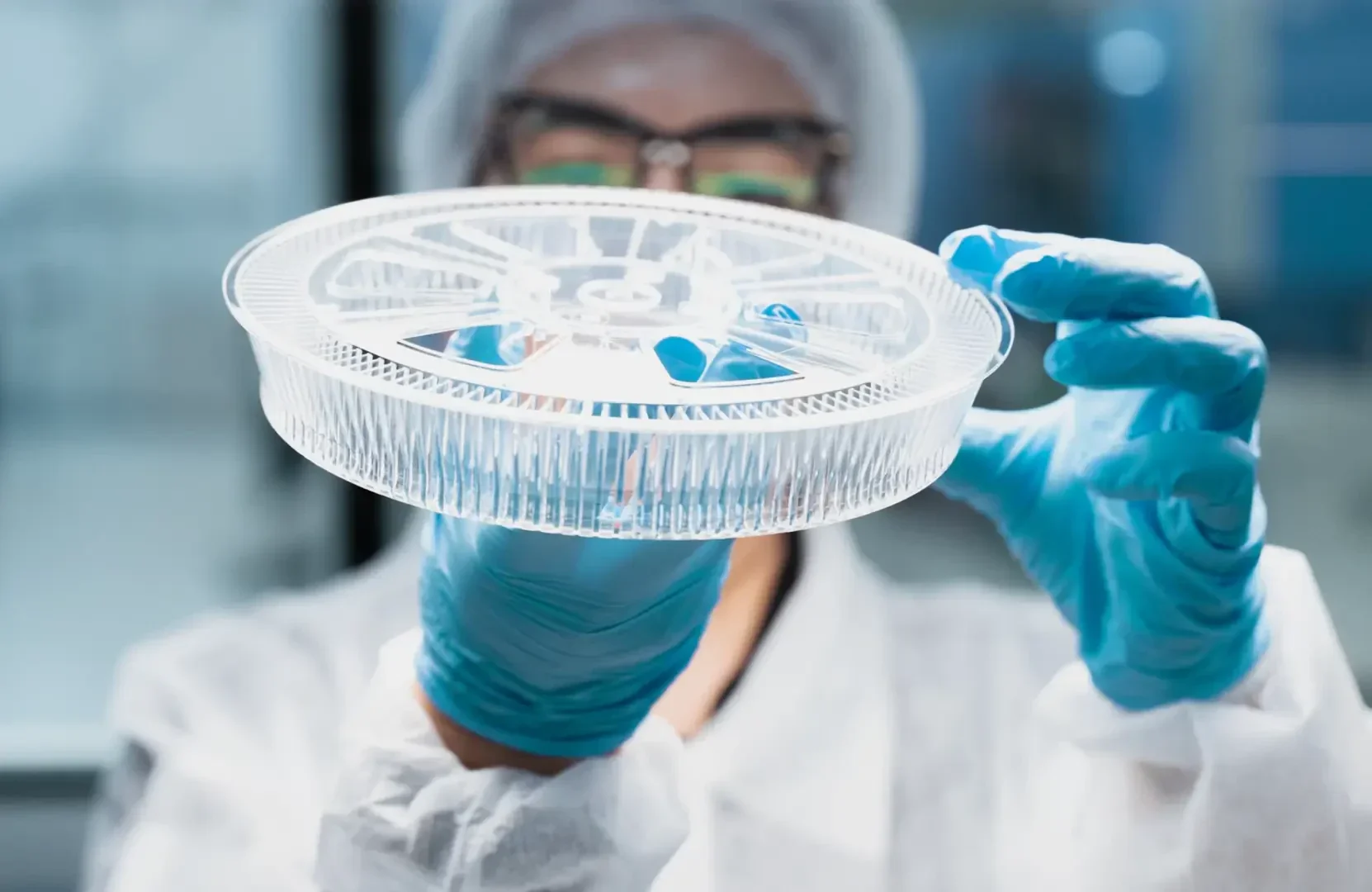 Ein Wissenschaftler, der einen Kittel, Handschuhe, eine Haube und eine Schutzbrille trägt, hält und untersucht in einem Labor eine große Petrischale aus durchsichtigem Kunststoff oder ein Probentablett.